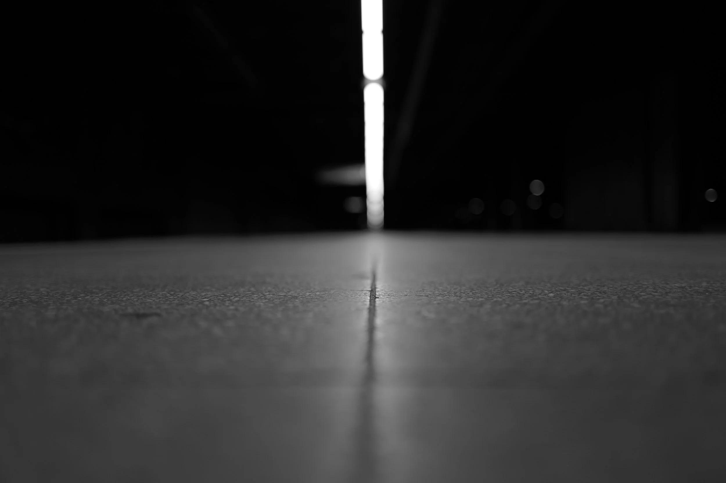 Photo en noir et blanc d'un sol avec une ligne droite centrale menant vers une source de lumière mystérieuse au fond, dans un environnement sombre et indéfini.