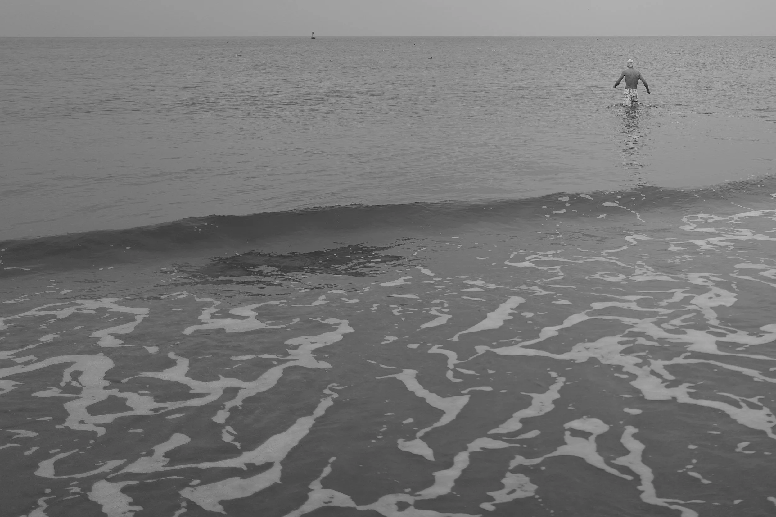 Un homme nage dans la mer, vue de derrière, avec de petites vagues jusqu'à lui, en noir et blanc.