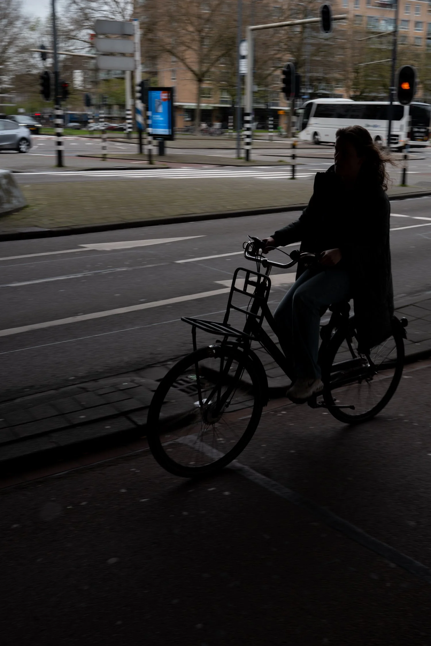 Une personne à vélo dans un espace urbain, silhouette sombre, avec un fond de rue avec des voitures et des bus, arbres et bâtiments en arrière-plan.