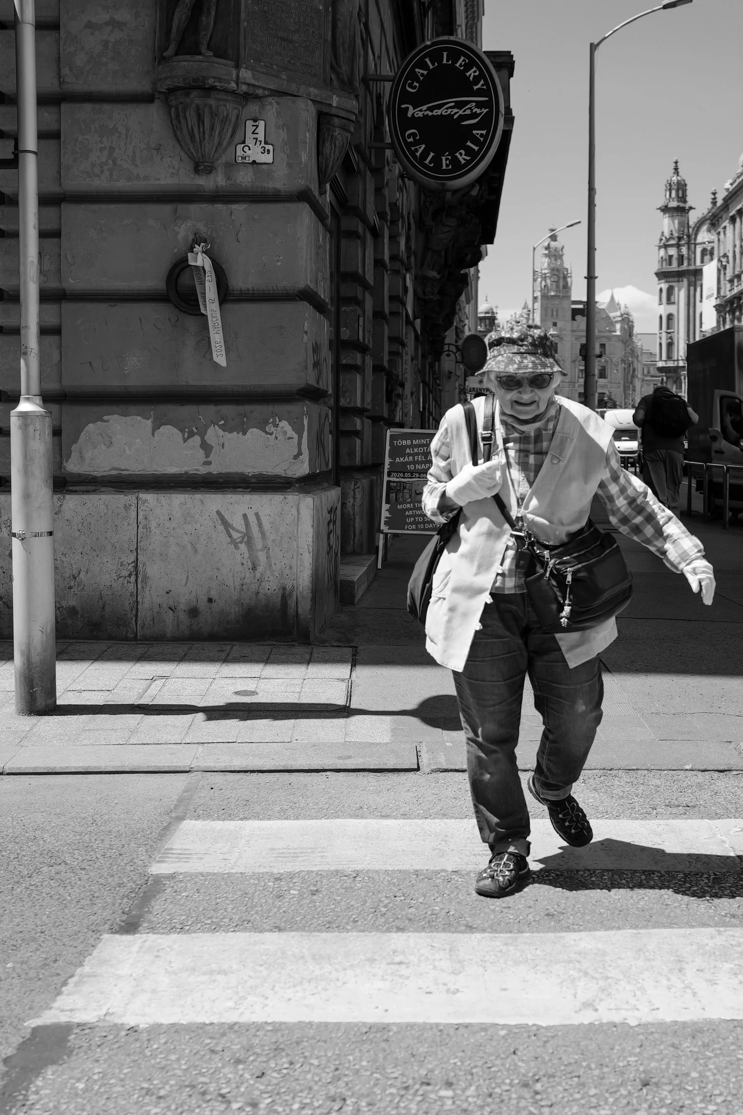 Une personne âgée, portant des lunettes et un chapeau, traverse un passage piéton dans une ville, avec des bâtiments historiques en arrière-plan.