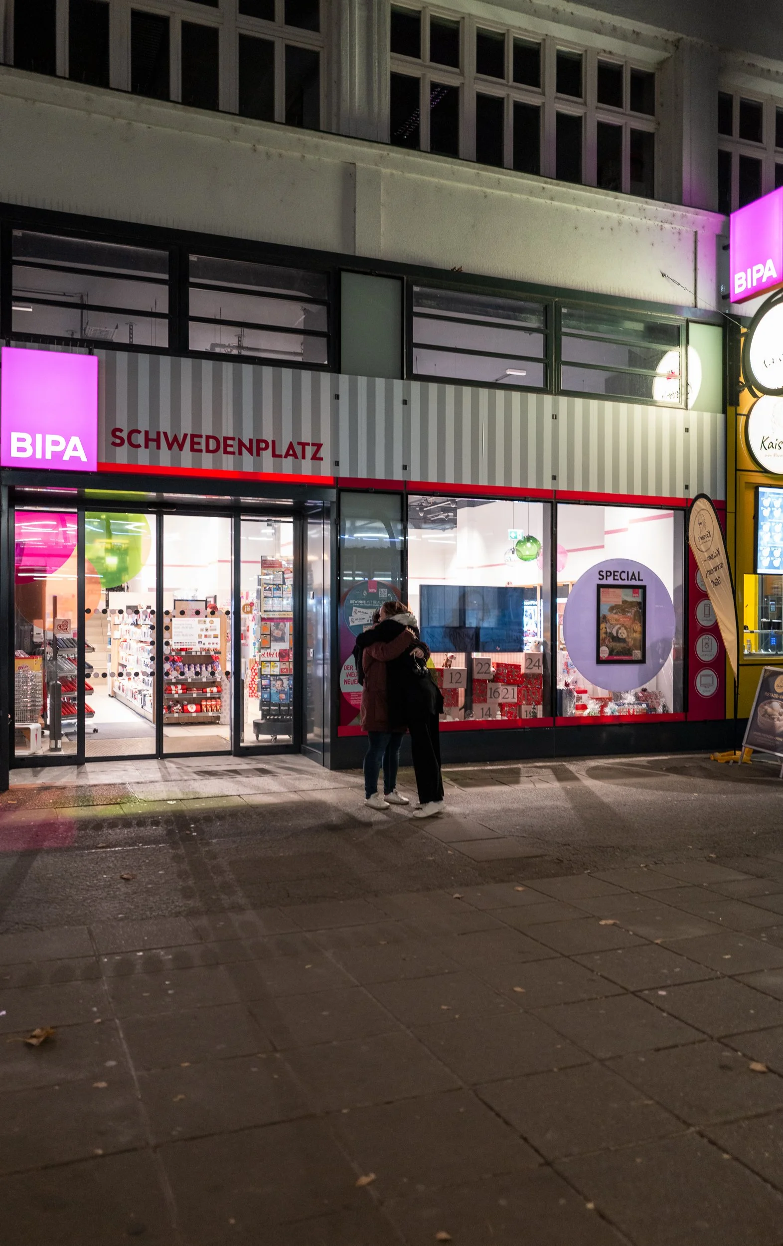 Deux personnes en hiver se tiennent devant une boutique nommée 'BIPA' à Schwedenplatz, en Autriche, la nuit, sous un ciel sombre, avec des fenêtres donnant sur des produits à l’intérieur et un éclairage lumineux.