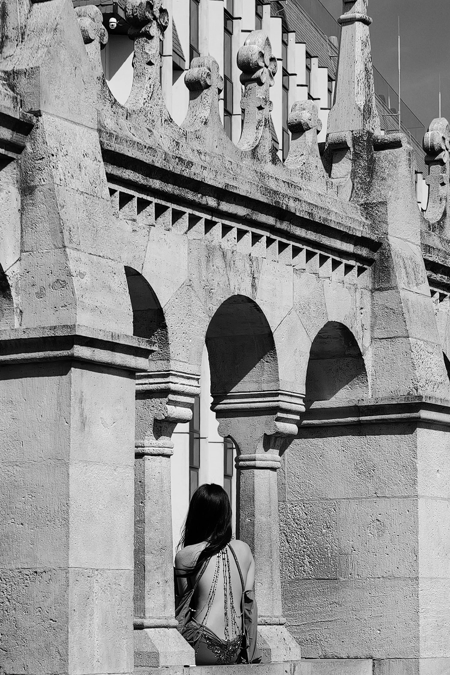 Une femme avec un sac, vue de dos, se tient devant une architecture en pierre ornée de motifs en forme de crânes et de flèches, avec un bâtiment moderne à fenêtres en arrière-plan.
