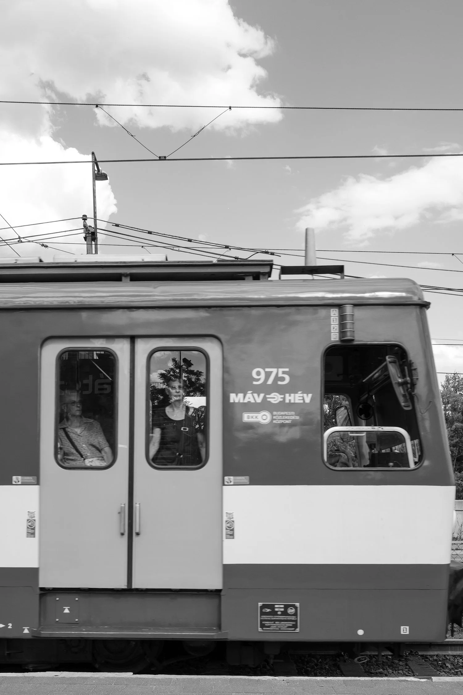 Photo en noir et blanc d'un tramway avec des passagers à l'intérieur et des lignes électriques au-dessus, sous un ciel partiellement nuageux.