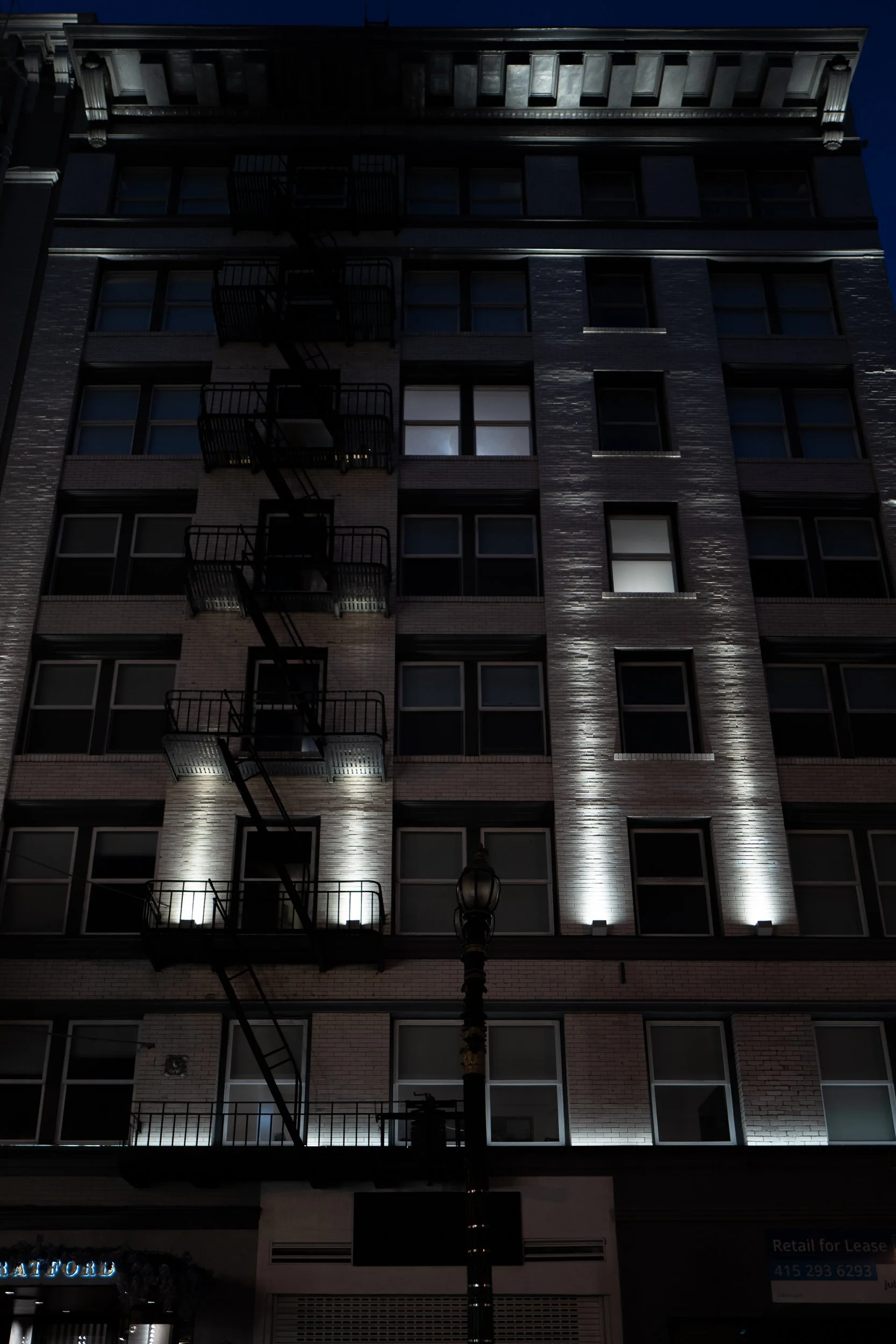 Un bâtiment résidentiel de plusieurs étages éclairé la nuit, avec une cheminée en métal noir, des fenêtres, et un lampadaire devant l'immeuble.