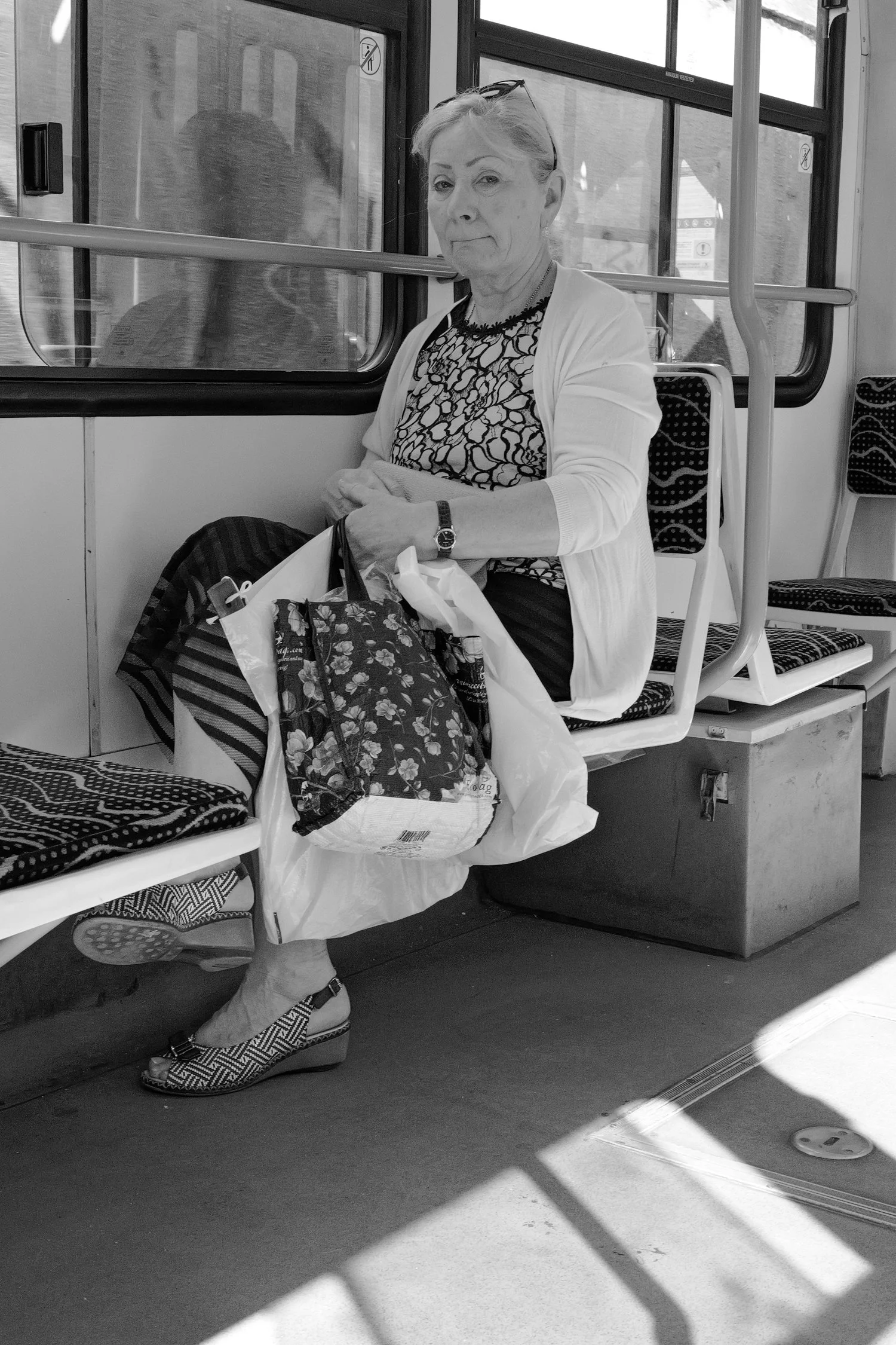 Une femme âgée assise dans un bus, portant une robe à motifs et une veste blanche, portant une montre et des baskets à motifs. Elle tient un sac de shopping sur ses genoux. Elle regarde vers l'appareil photo.