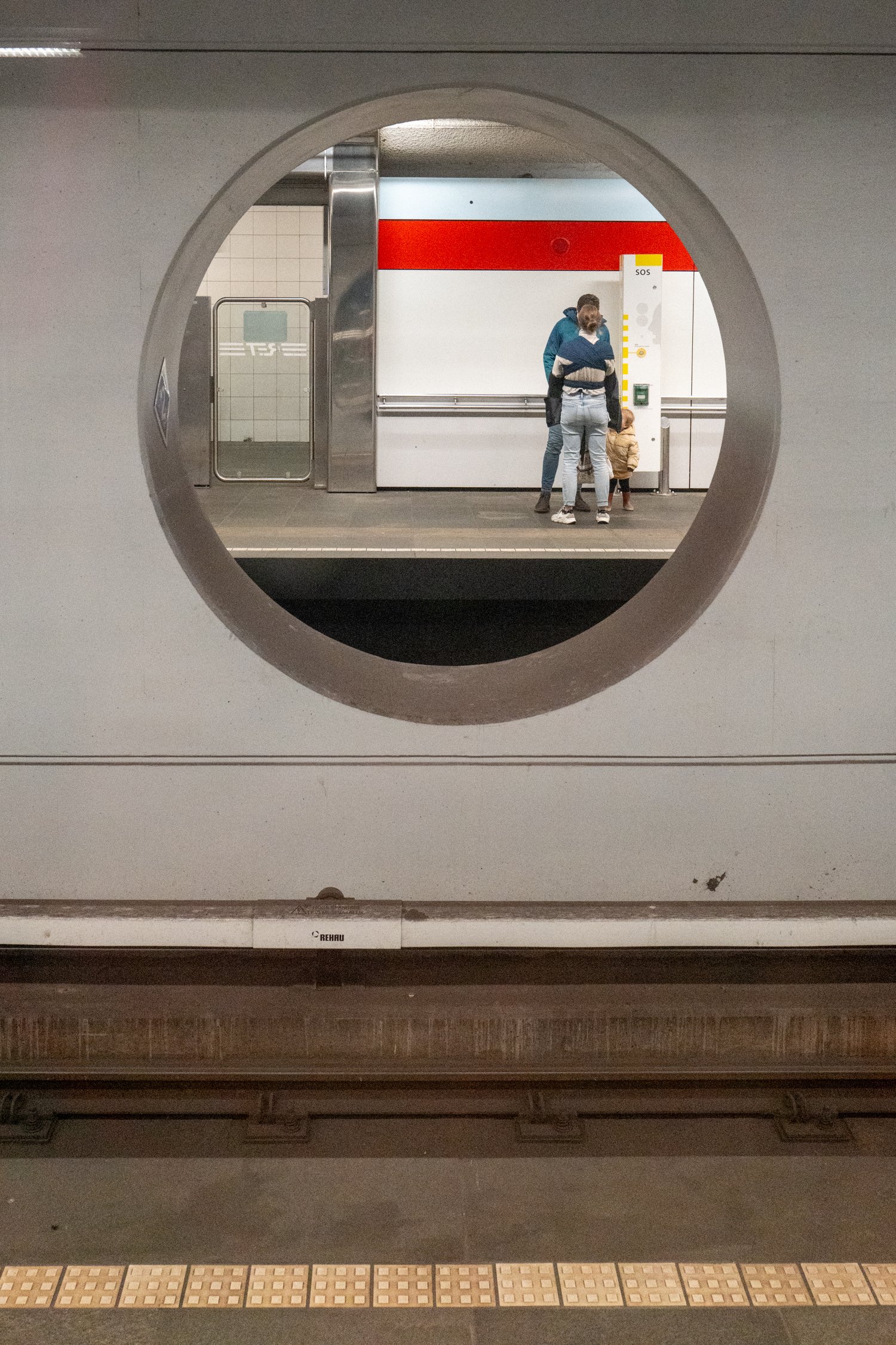 Vue d'une station de métro à travers une fenêtre circulaire. On voit des personnes attendant sur le quai, un panneau SOS, des carreaux sur le mur et des éléments métalliques.