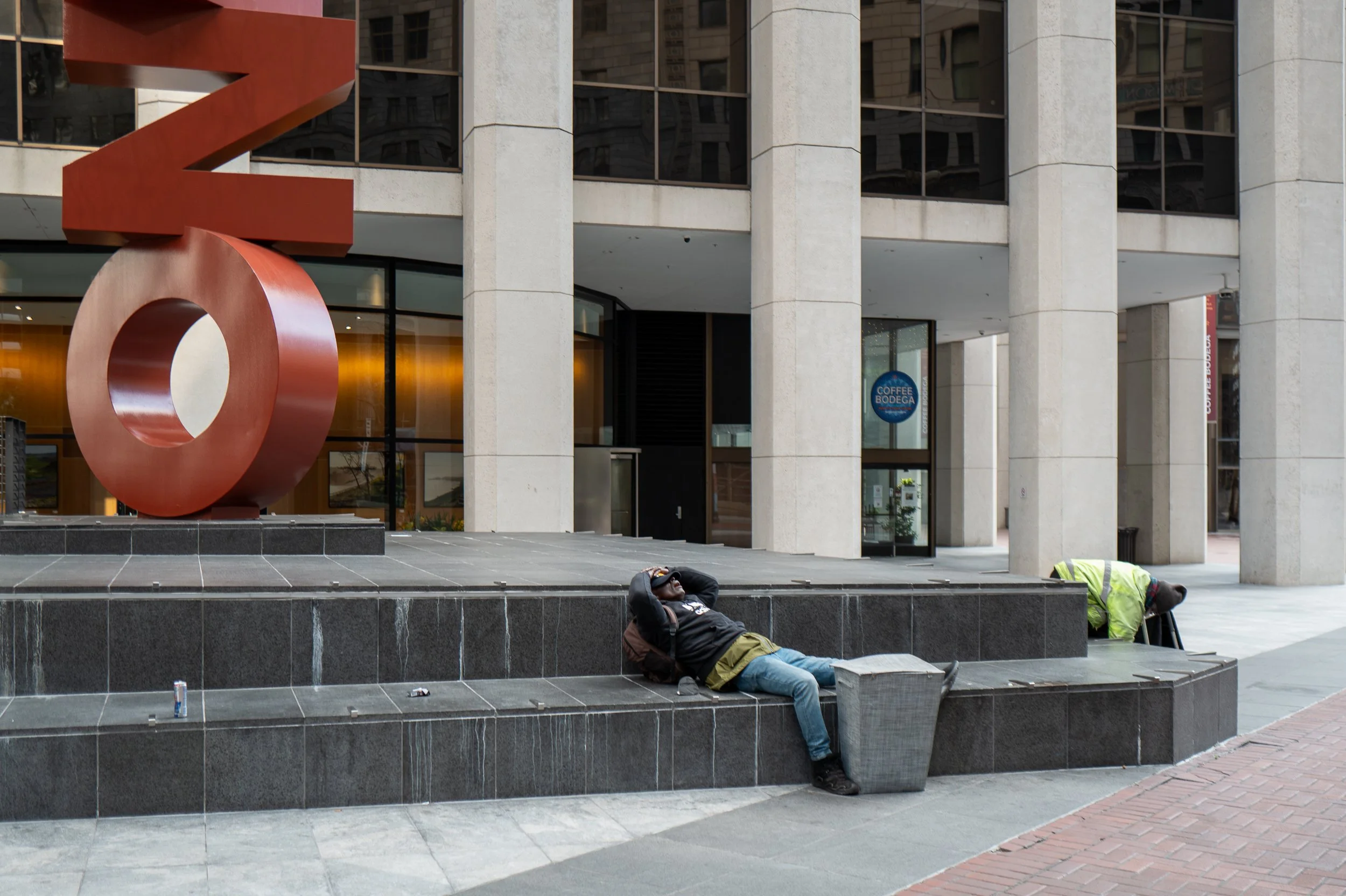 Deux personnes dorment sur un banc dans une zone urbaine devant un bâtiment moderne avec une sculpture rouge
