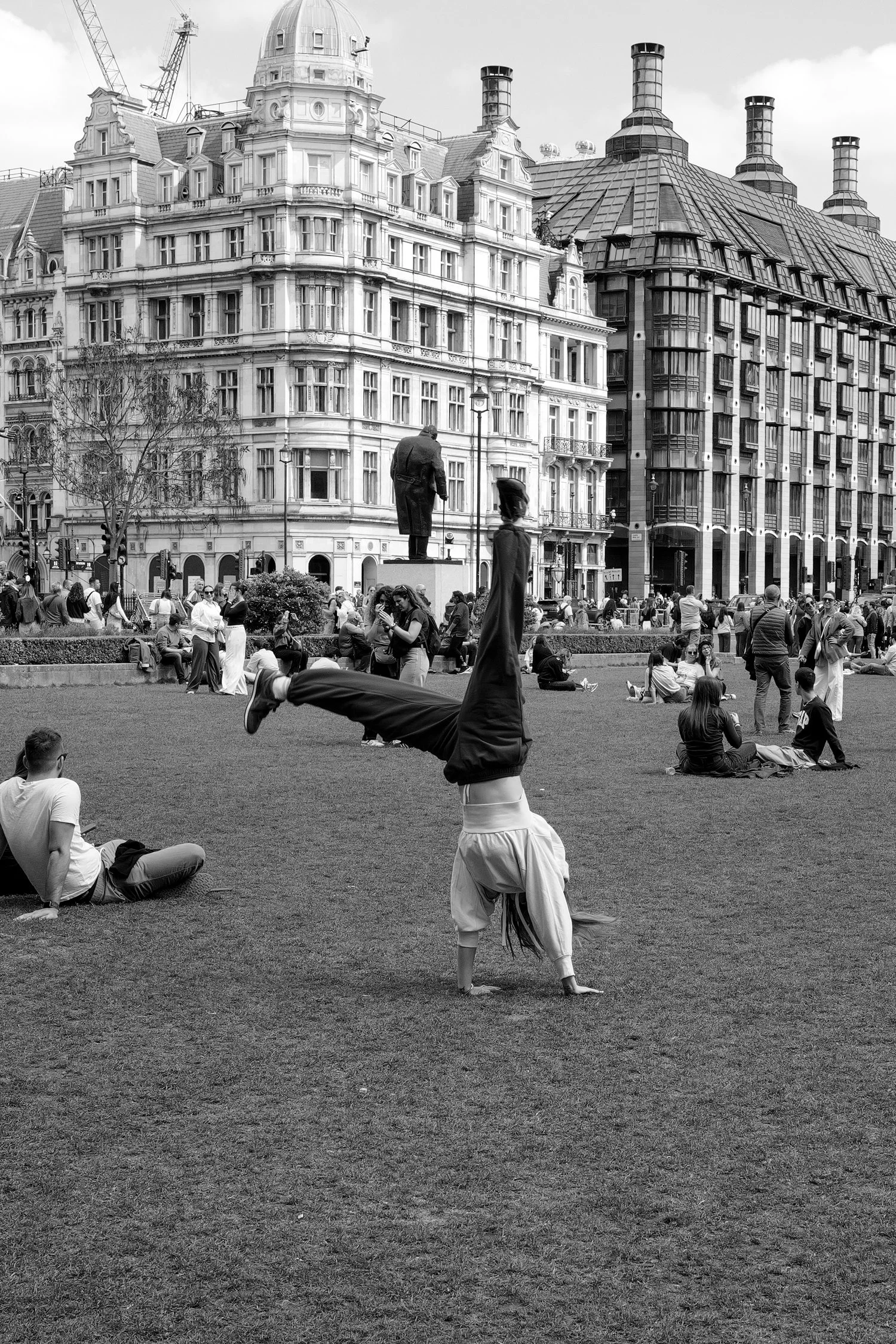 Une place animée avec des personnes assises, debout ou se pratiquant la gymnastique, des bâtiments historiques et modernes en arrière-plan, dont un établissement avec plusieurs cheminées.