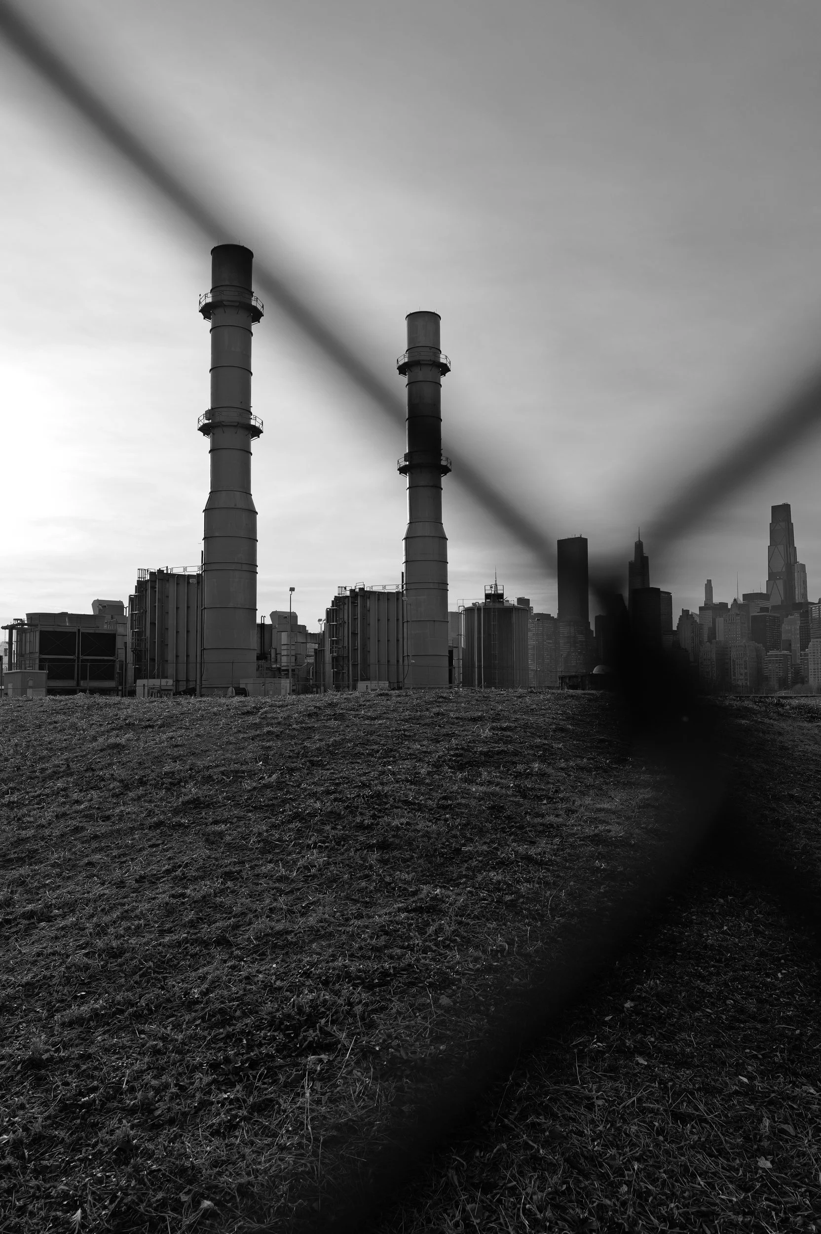 Une usine avec deux grandes cheminée dans une ville moderniste, vue à travers une clôture en fil de fer, en noir et blanc.