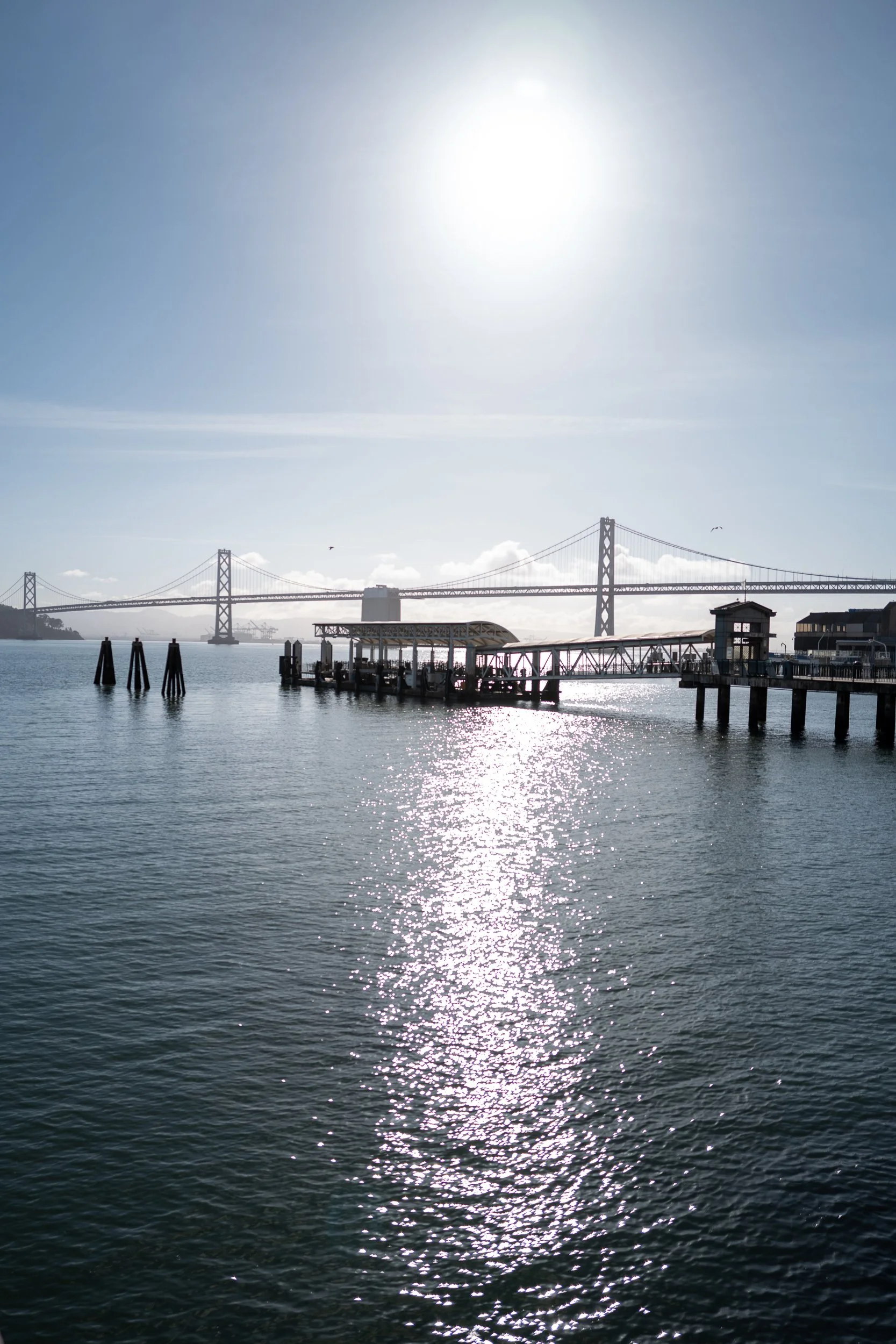 Vue d'un quai au bord de l'eau avec un pont en arrière-plan, sous un ciel ensoleillé.