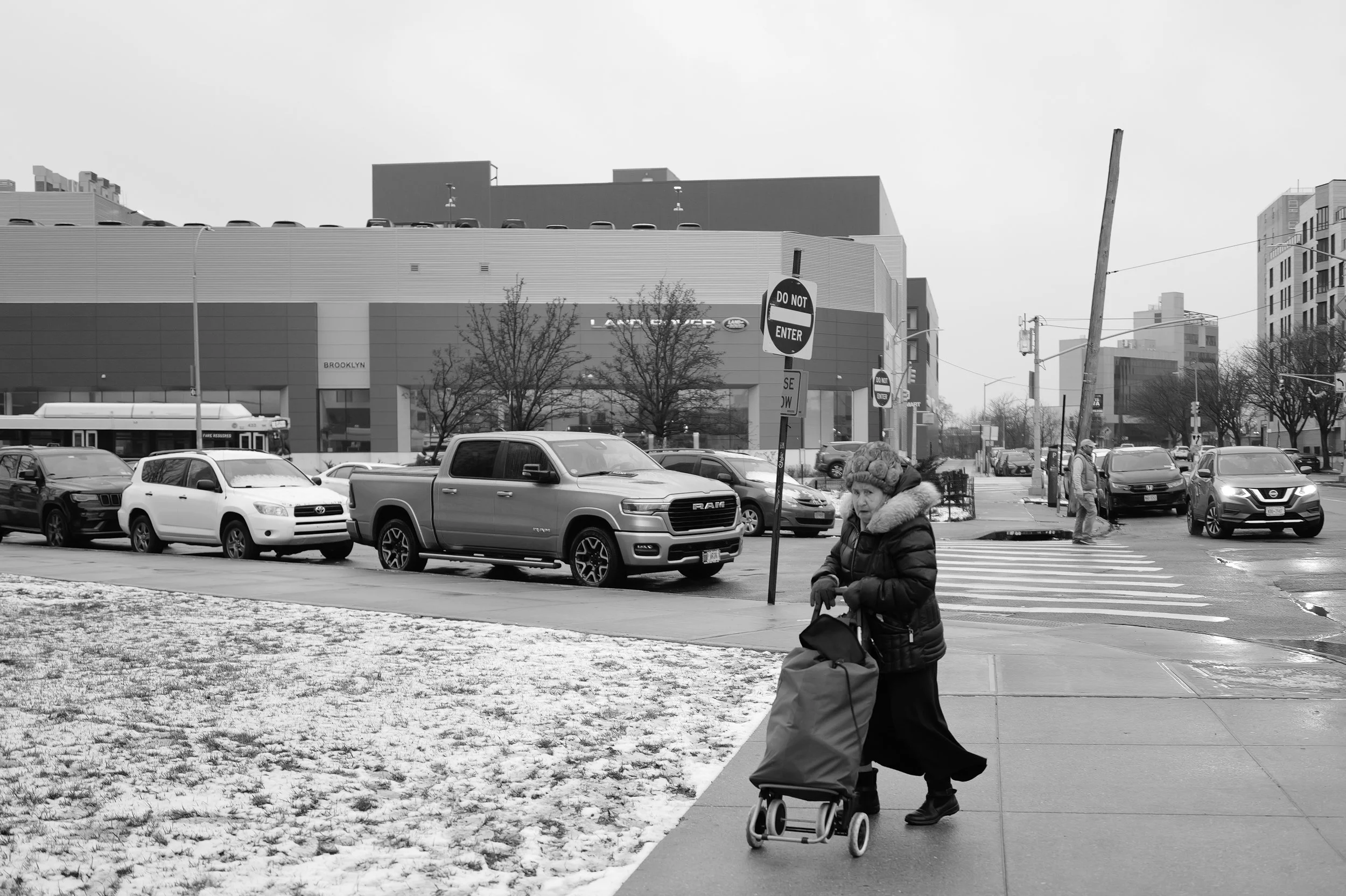 Une femme âgée portant un manteau avec une capuche en fourrure pousse un caddie sur le trottoir, devant un parking avec plusieurs voitures. Il y a des arbres sans feuilles, un bâtiment commercial, et des panneaux de signalisation dont un « Interdire 