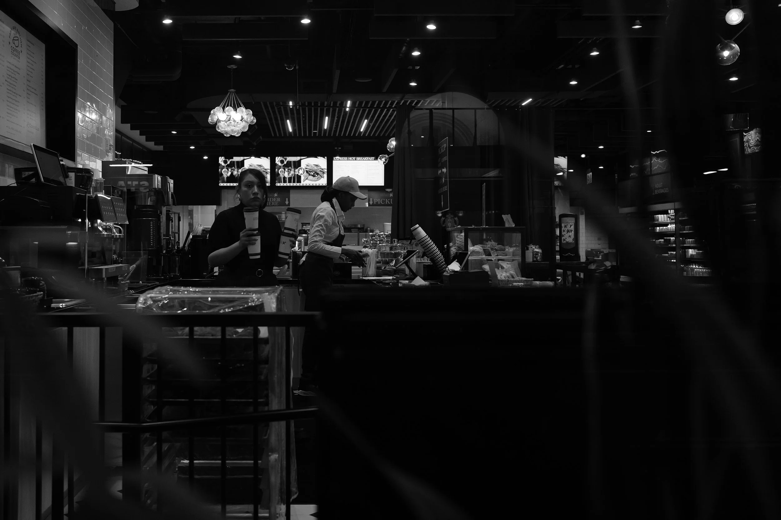 Deux employés dans un café ou une boutique, une portant une casquette et utilisant le terminal, l'autre tenant un grand verre, avec des écrans au plafond et des rayons de produits en arrière-plan.