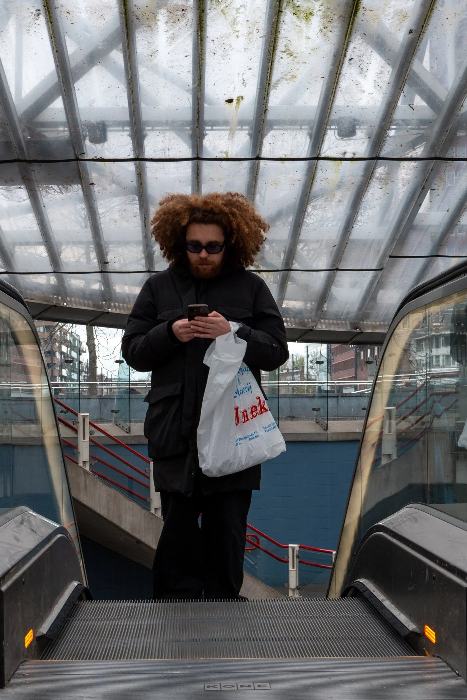 Une personne avec des cheveux bouclés et roux portant des lunettes de soleil, tenant un téléphone portable et un sac en plastique blanc, se trouve en haut d'une escalator dans une station de métro ou un centre commercial, vue de dessous.