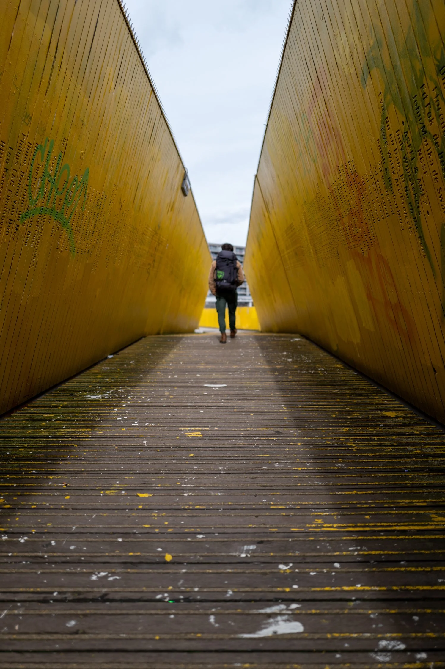Un homme avec un sac à dos marche entre deux murs en bois peint en jaune, avec un ciel gris en arrière-plan.