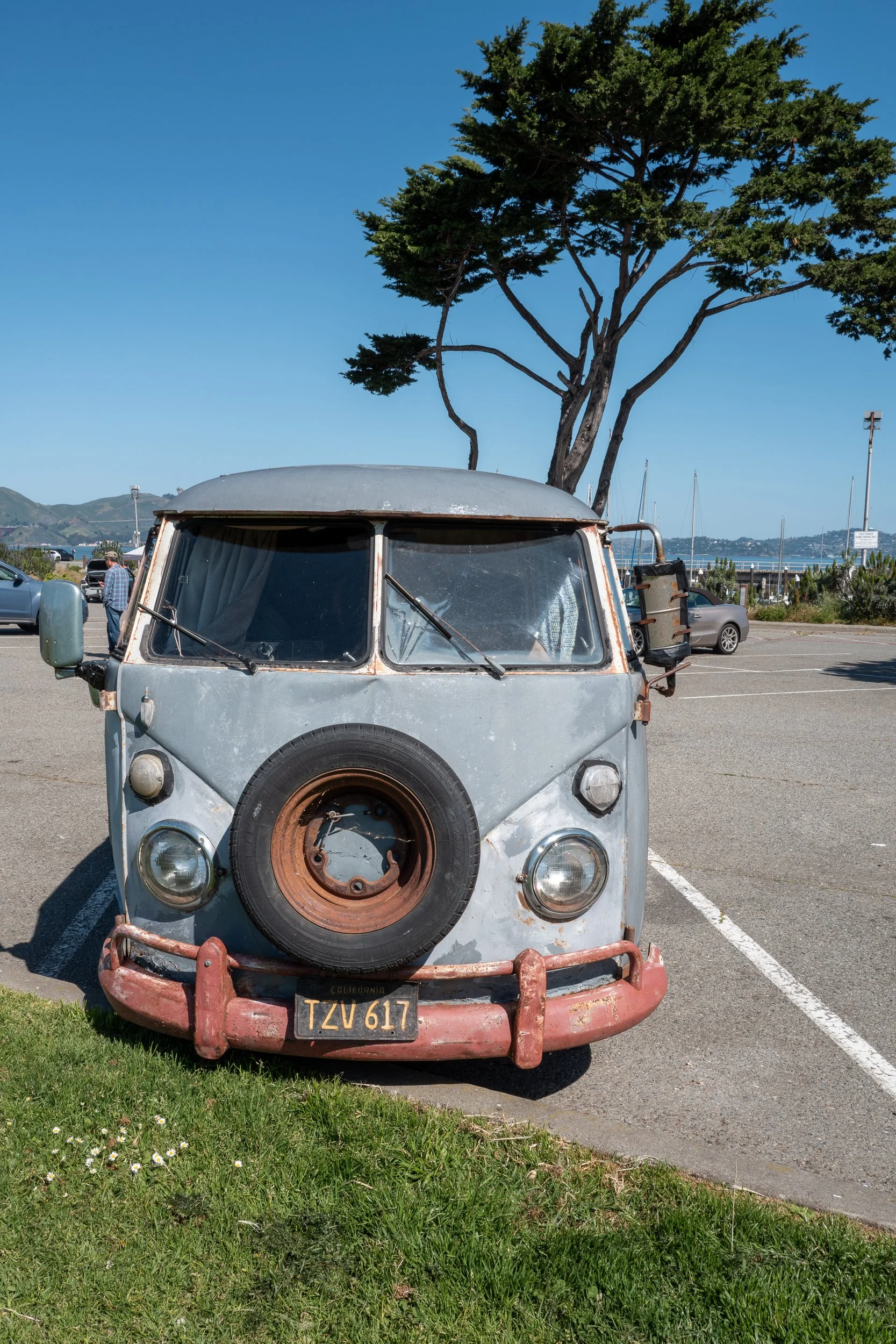 Une vieille camionnette vintage en métal gris avec une roue de secours en bois à l'avant, stationnée sur une place de parking près de la mer. Arbre grand et feuillu derrière, ciel bleu clair.