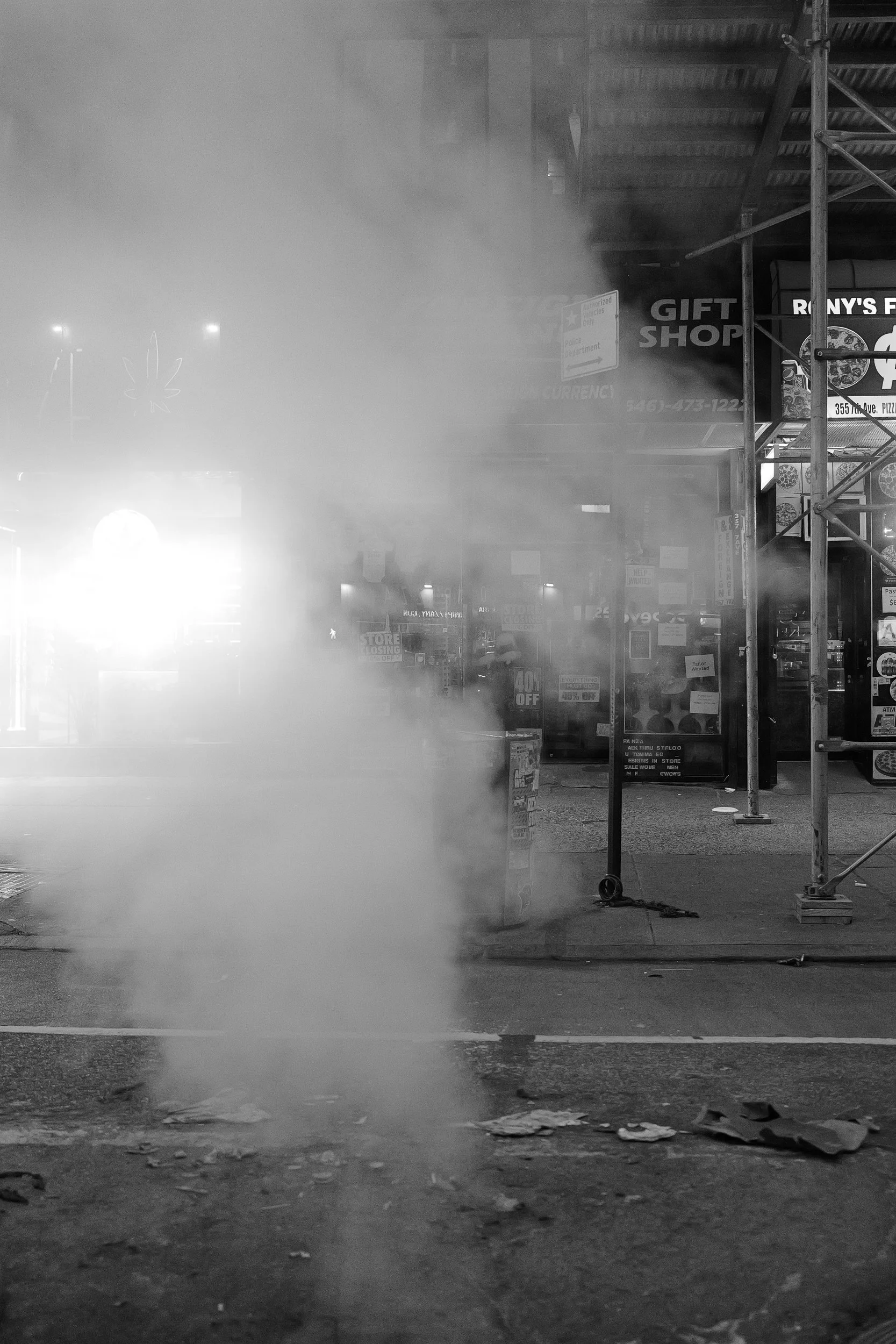 Une rue nocturne avec de la fumée ou de la vapeur s'échappant au sol, et des enseignes lumineuses floues en arrière-plan, dont une indiquant un magasin de cadeaux.