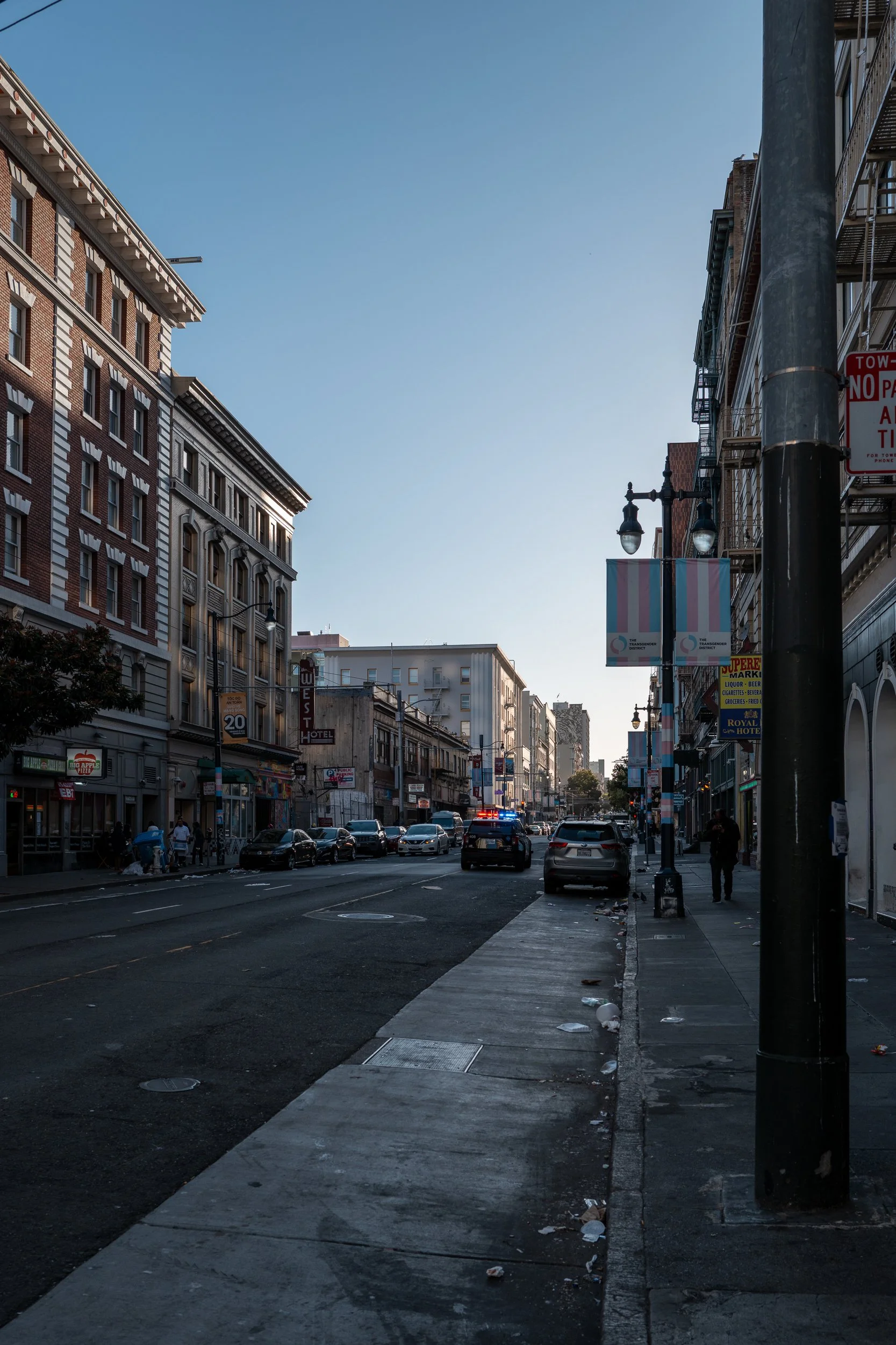 Une rue urbaine avec des bâtiments anciens, des voitures garées et des passants, sous un ciel clair au crépuscule ou à l'aube.