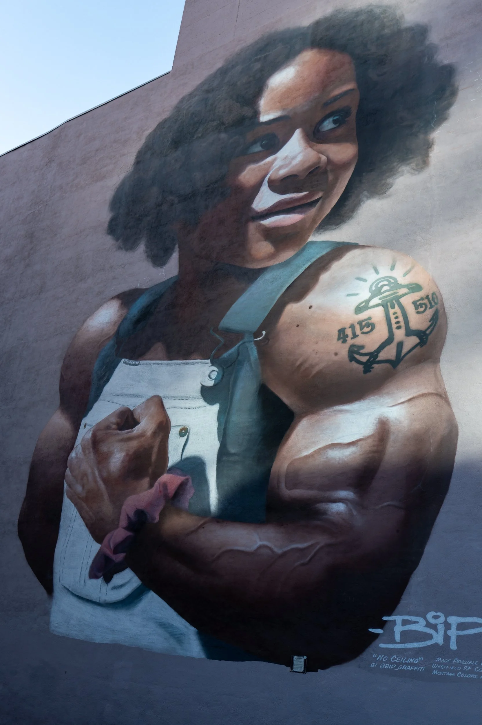 Une fresque murale représentant une femme afro-américaine souriante avec des cheveux bouclés, portant un pantalon en jean et levant son poing, avec un tatouage d'ancre sur son bras.