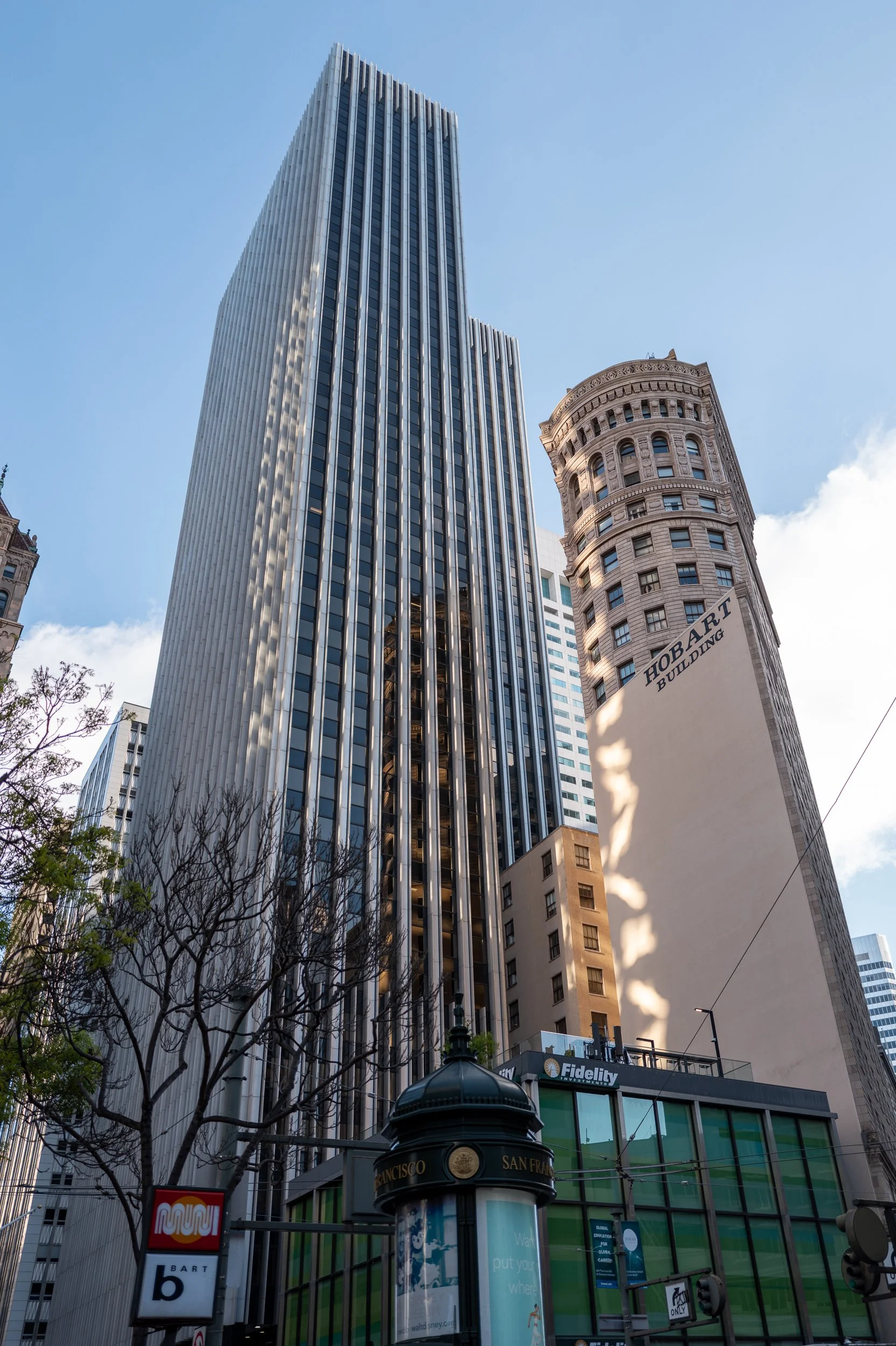 Vue sur des gratte-ciel urbains avec un ciel bleu et quelques nuages, incluant un bâtiment moderne et un bâtiment historique avec l'inscription 'Hobart Building', avec des arbres en premier plan et un panneau 'Fidelity'.