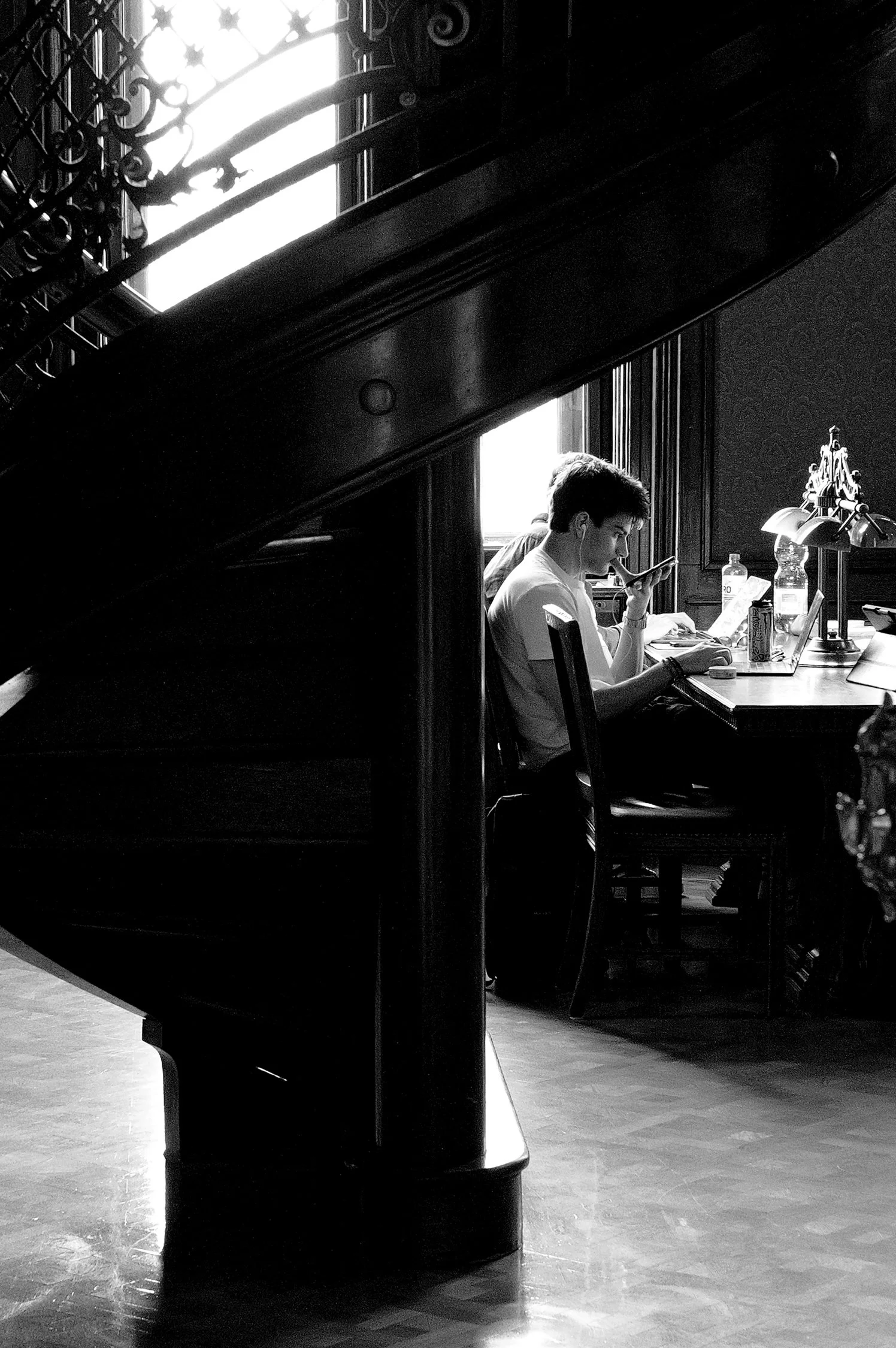 Un jeune homme assis à un bureau, avec des lampes de bureau, en regardant son téléphone, dans une pièce avec une grande fenêtre.