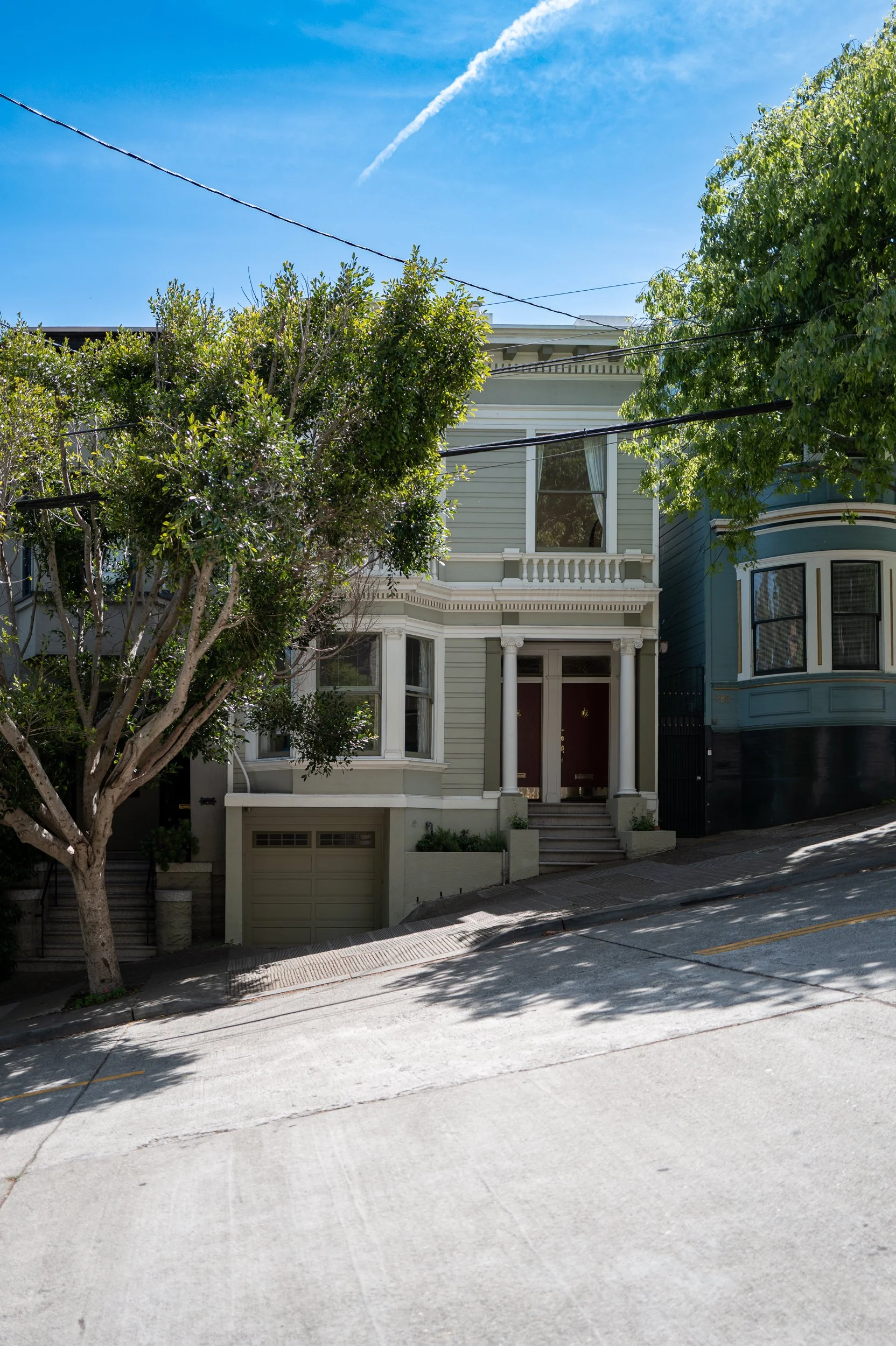 Maison victorienne à plusieurs étages avec un porche et deux colonnes, entourée de végétation, sur une rue en pente, sous un ciel bleu.