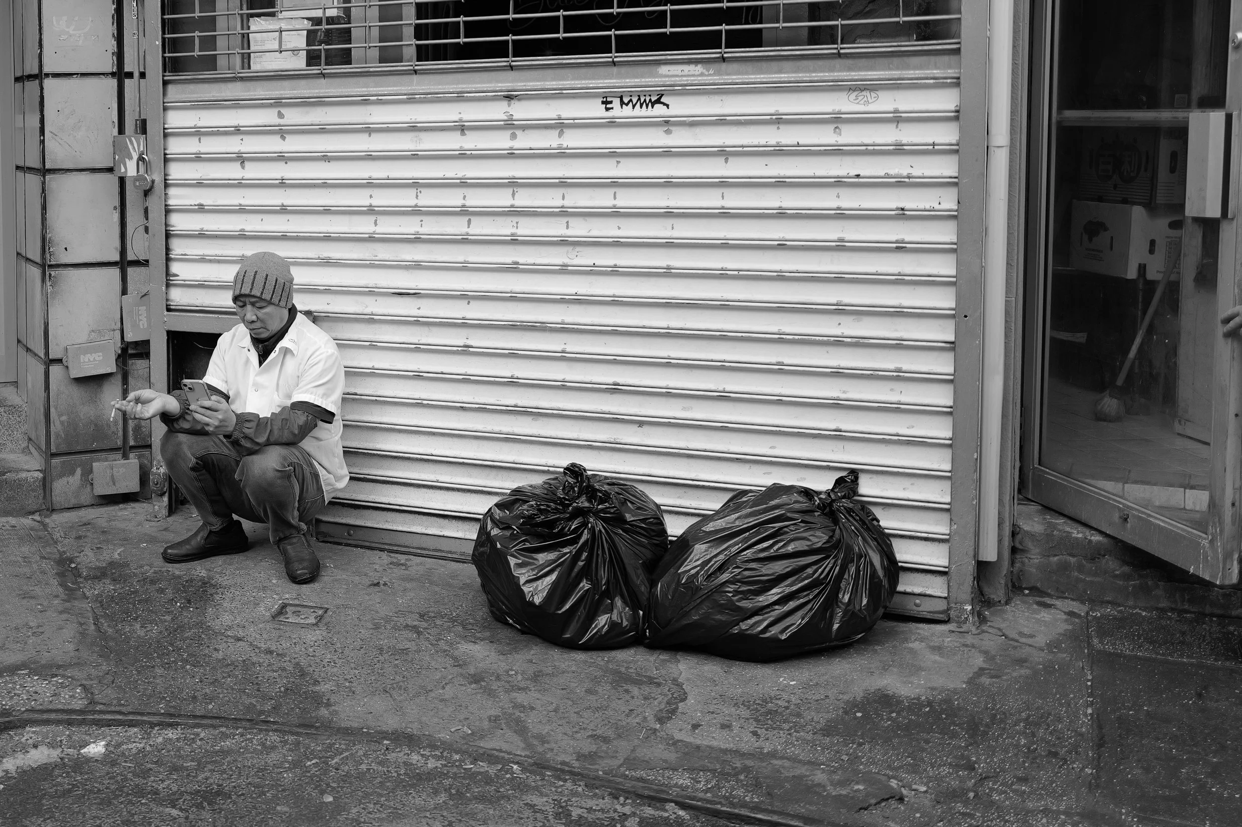Un homme assis par terre à côté de sacs poubelles noires, utilisant son téléphone, devant un rideau métallique fermé, dans une rue urbaine.