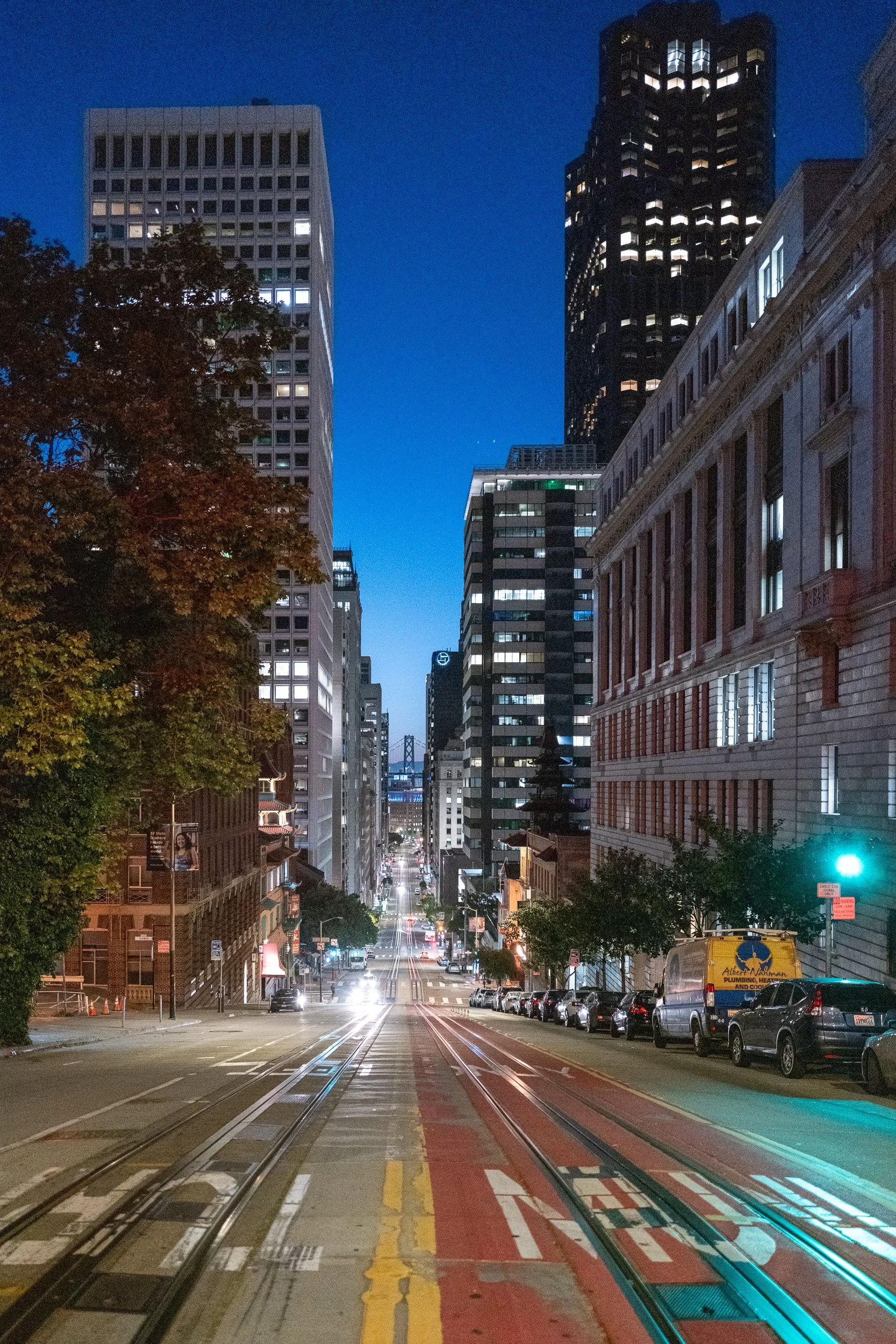 Une vue urbaine nocturne d'une rue de San Francisco avec des bâtiments modernes, des arbres et des rails de tramway, avec le pont du Bay Bridge visible au loin.