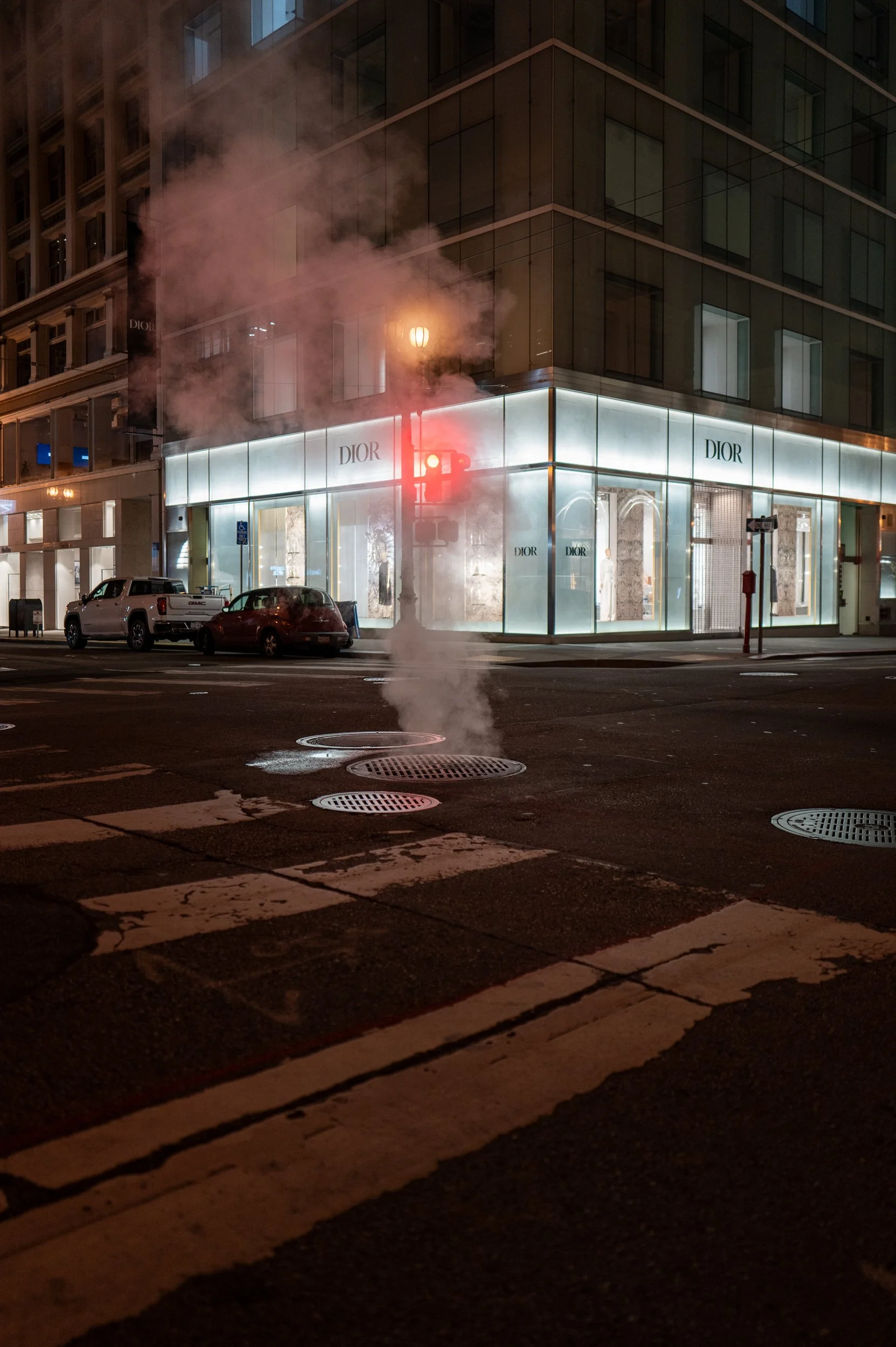 Une rue urbaine la nuit avec un bâtiment Dior illuminé. Il y a de la vapeur ou de la fumée s'échappant d'une bouche d'incendie au centre de la scène, ainsi que quelques voitures garées sur le côté de la rue.