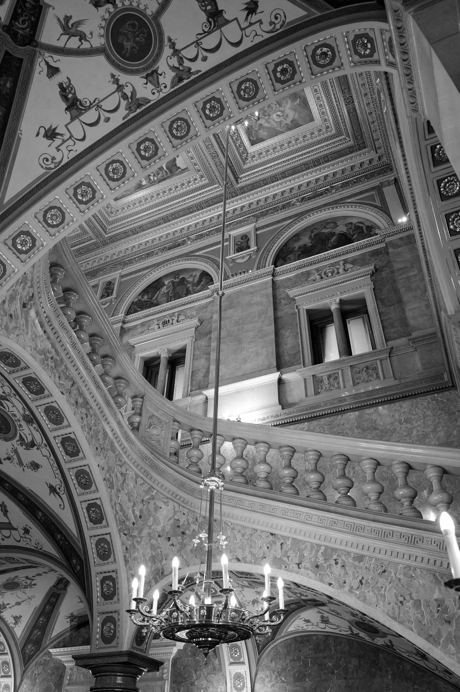 Intérieur somptueux d’un bâtiment historique avec des plafonds ornés, des fresques, une balustrade en pierre et un chandelier en métal avec des bougies.