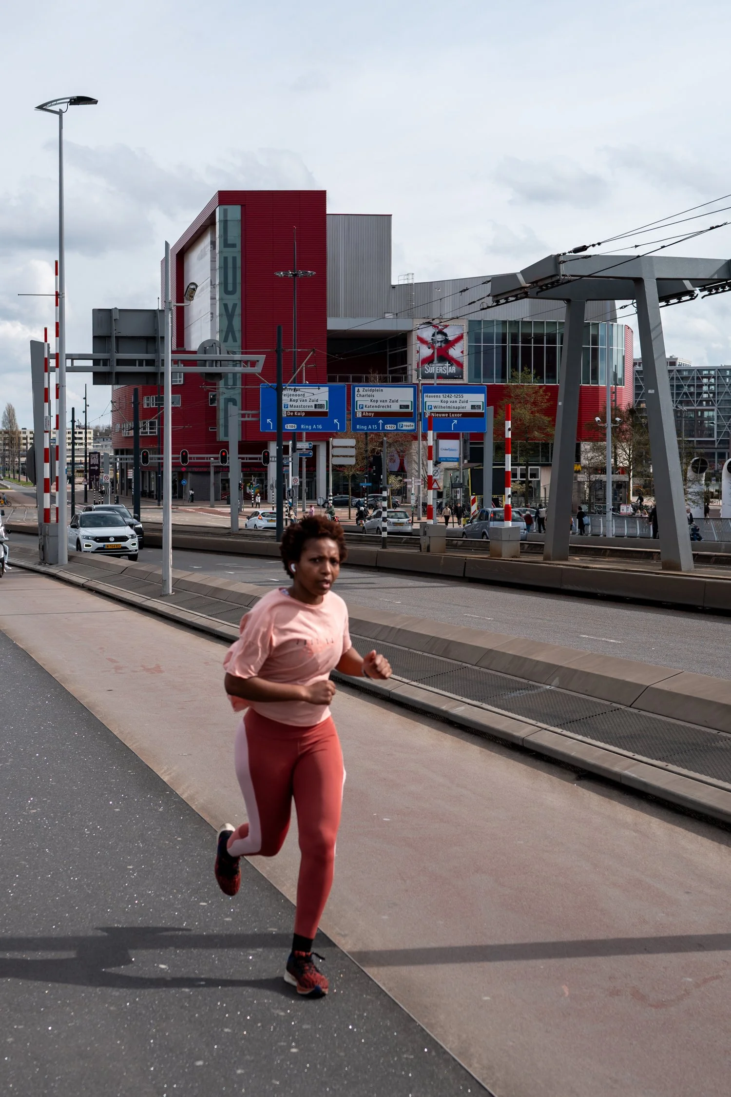 Une femme court en pleine rue, portant un ensemble de sport rose et rouge, avec des chaussures de course noires et rouges, avec un bâtiment moderne rouge et gris en arrière-plan et des panneaux de signalisation routière.