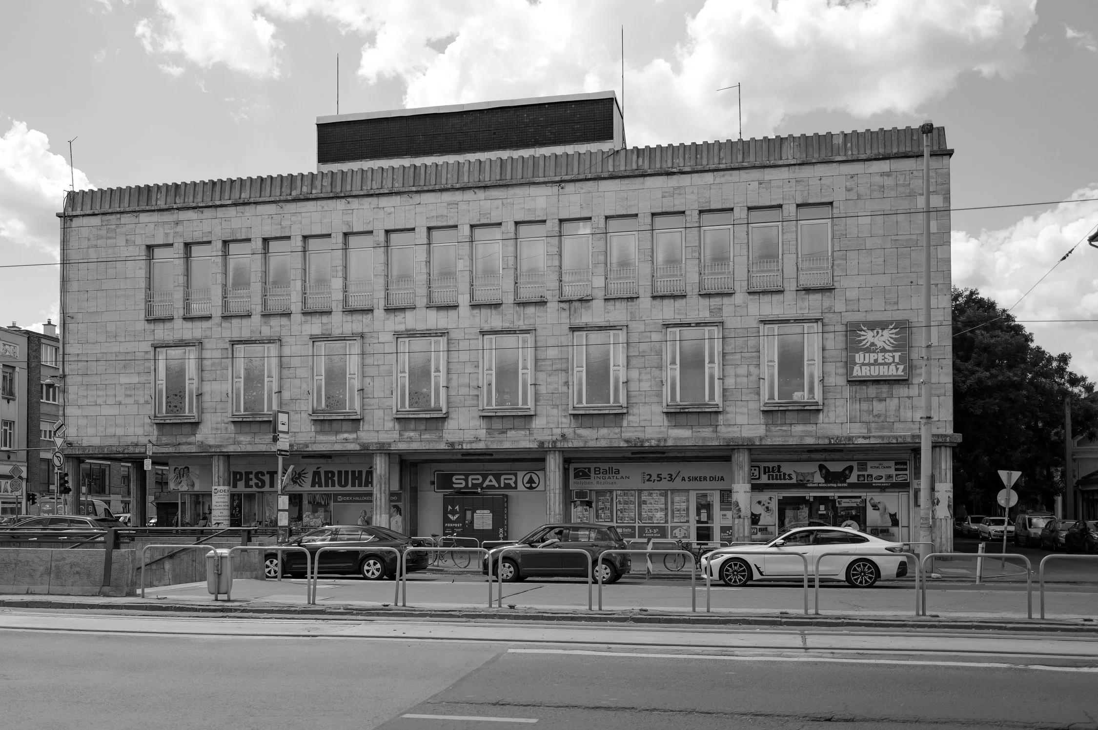 Un bâtiment moderne en pierre avec plusieurs fenêtres et un panneau publicitaire en bas. En premier plan, il y a une rue avec des voitures garées et des barres de sécurité le long du trottoir.