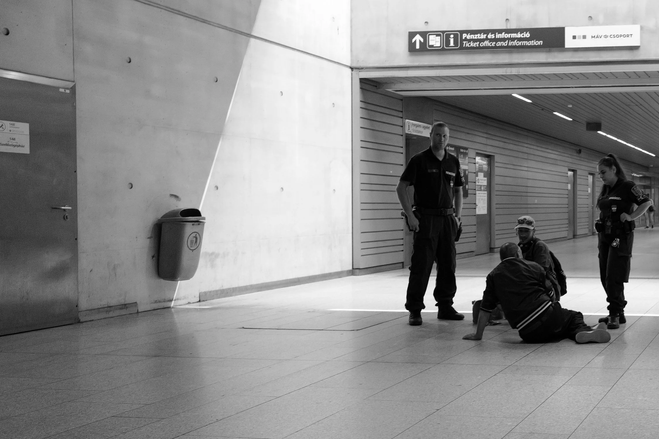 Deux agents de sécurité en uniforme discutent, assis par terre dans une gare ou un aéroport, avec d'autres personnes assises à proximité et une signalisation indiquant le bureau des billets et l'information.