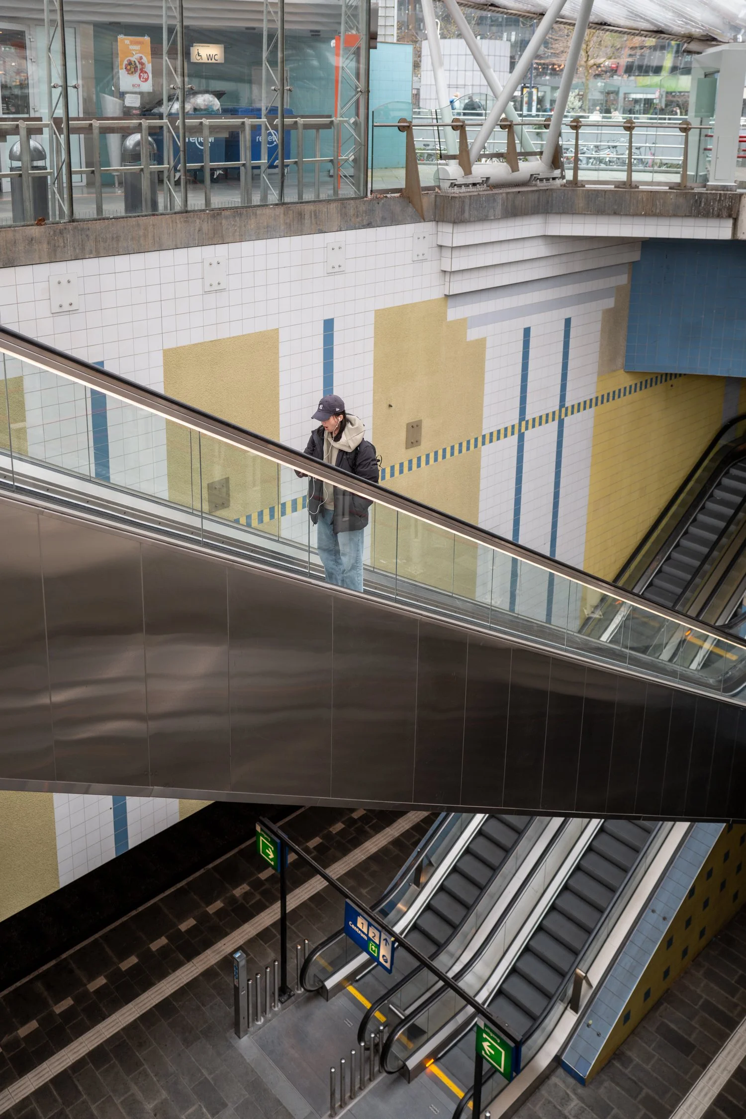 Une scène d'intérieur d'une station de métro ou d'un centre commercial avec des escalators, un mur décoratif, une personne portant un manteau noir, un chapeau, et des écouteurs. On voit aussi un panneau d'évacuation vert et des escaliers mécaniques.