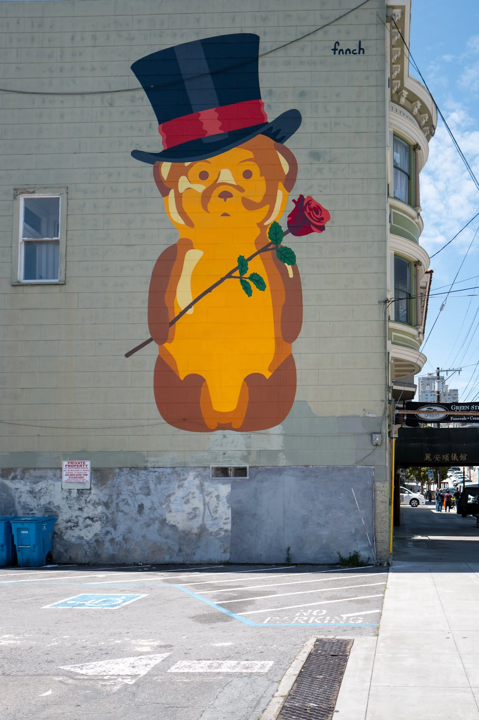 Mural représentant un ourson avec un chapeau haut de forme et une rose dans la bouche, en style cartoon, peint sur un mur de bâtiment, dans une zone urbaine.