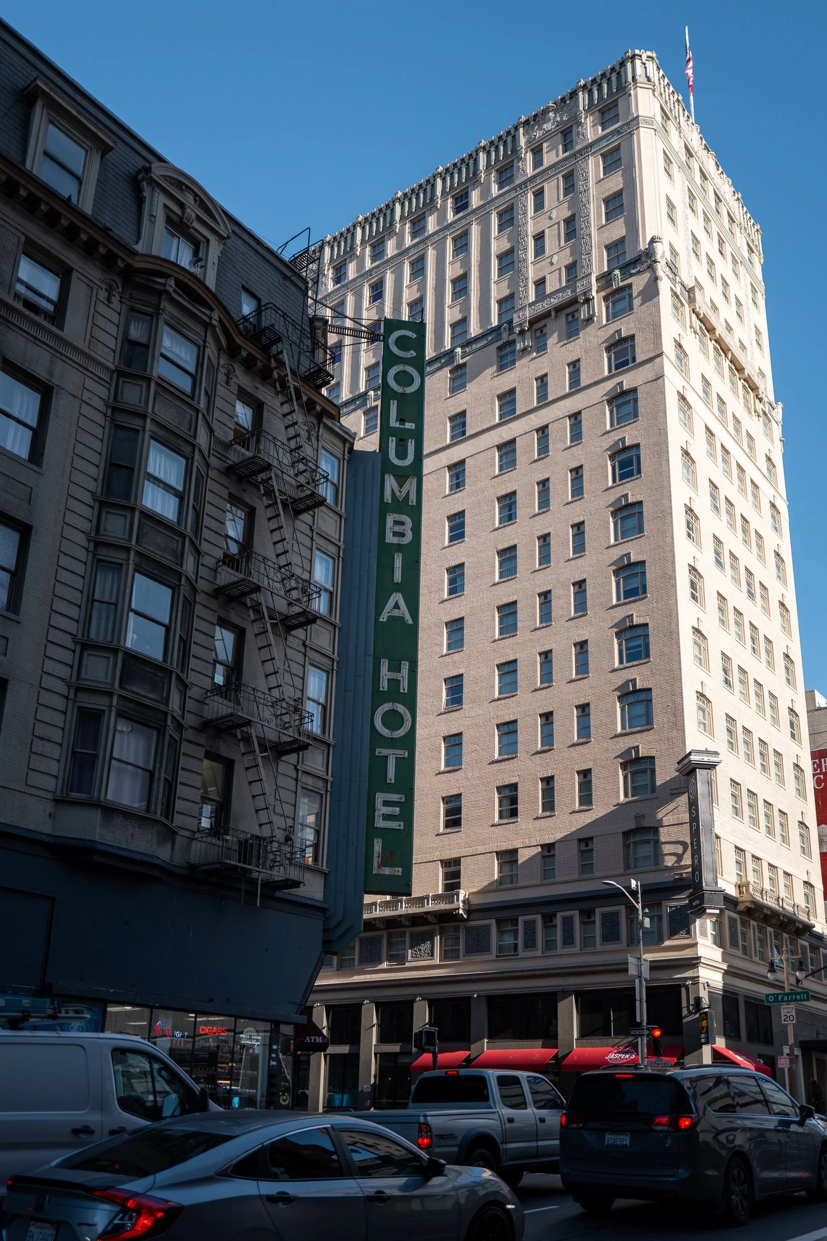 Vue urbaine montrant un bâtiment avec un grand signe vert indiquant 'COLUMBIA HOTEL' et plusieurs voitures en circulation à l'avant.