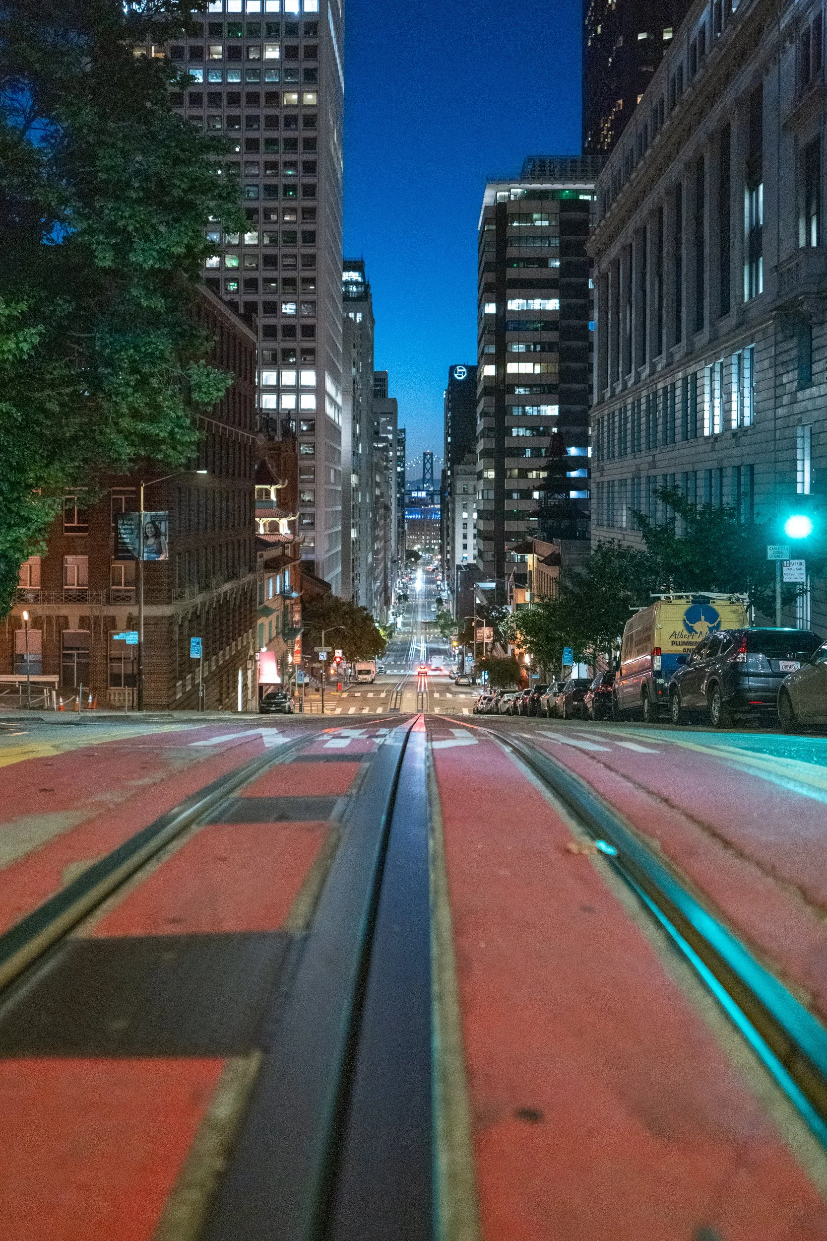 Vue sur une rue de ville la nuit, avec des bâtiments modernes, des voitures garées, et une ligne de tramway au centre