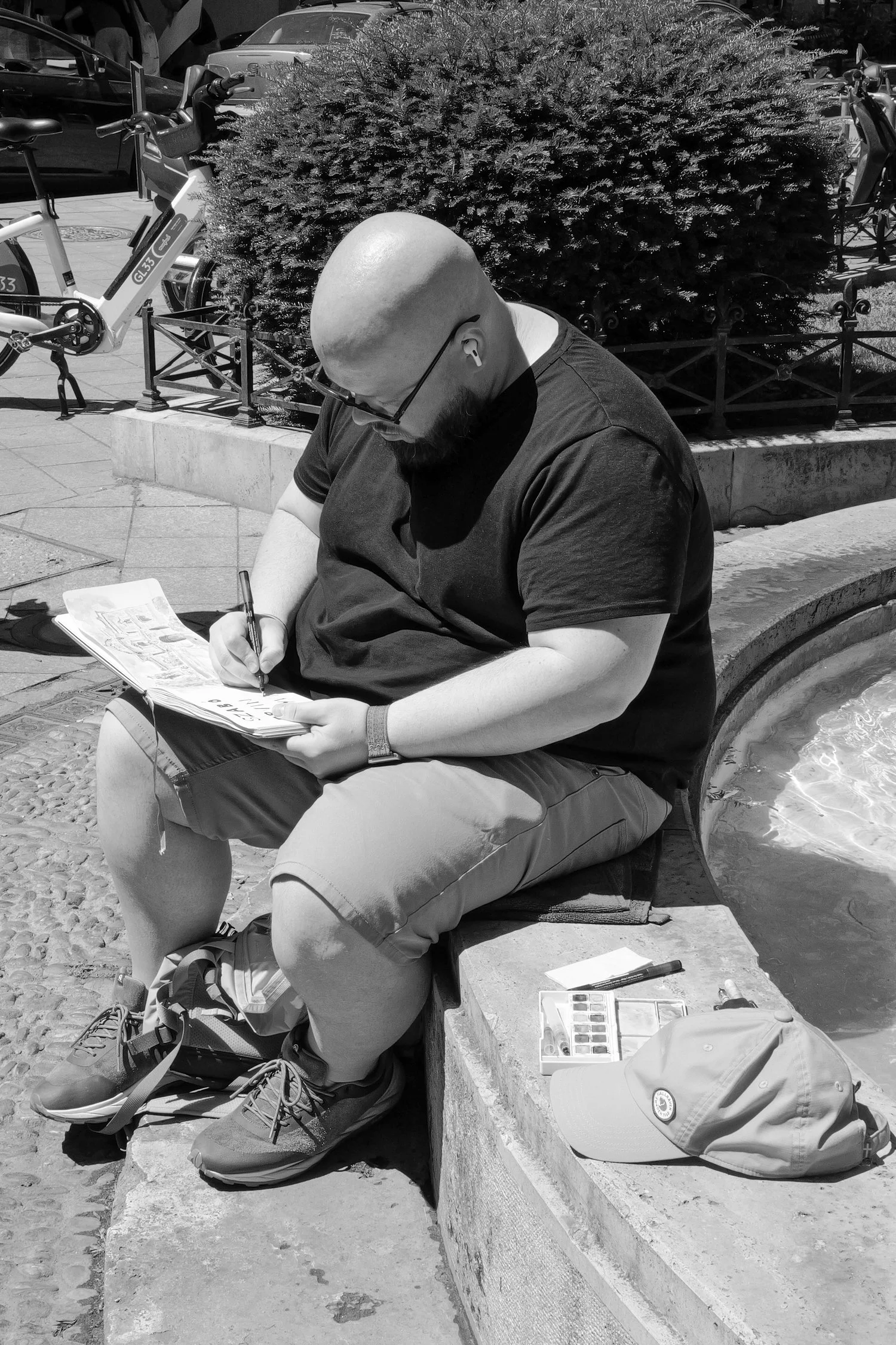 Un homme chauve portant des lunettes, assis sur un banc, écrit dans un journal, avec une casquette posée à côté. Il porte des écouteurs et des vêtements décontractés, dans un environnement urbain avec des vélos, des voitures et des arbustes en arrièr