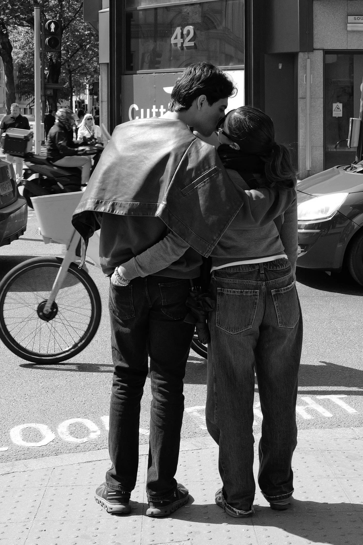 Deux jeunes personnes se tiennent très proches, se faisant un bisou sur la bouche dans une rue urbaine en plein jour. L’un porte une veste en cuir et l’autre un sweat, tous deux en jeans. Un vélo passe devant eux et il y a plusieurs passants en arriè