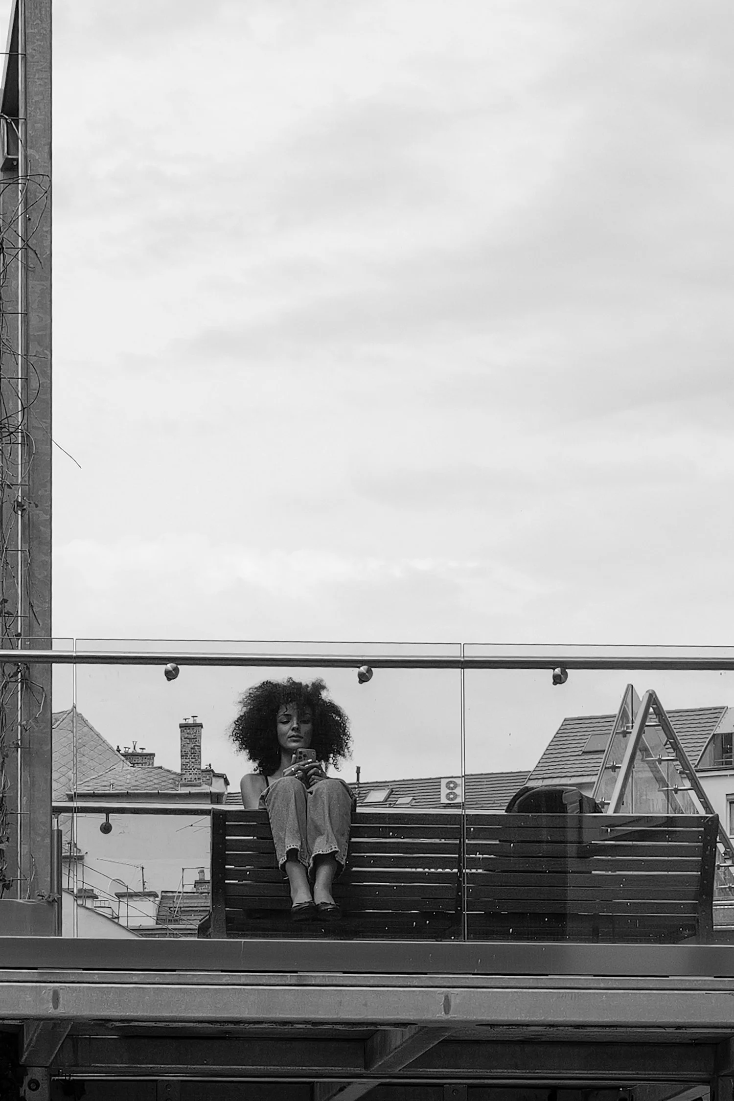 Une jeune femme avec des cheveux bouclés assise sur un banc en pleine ville, prenant une selfie, avec des bâtiments et un ciel nuageux en arrière-plan.