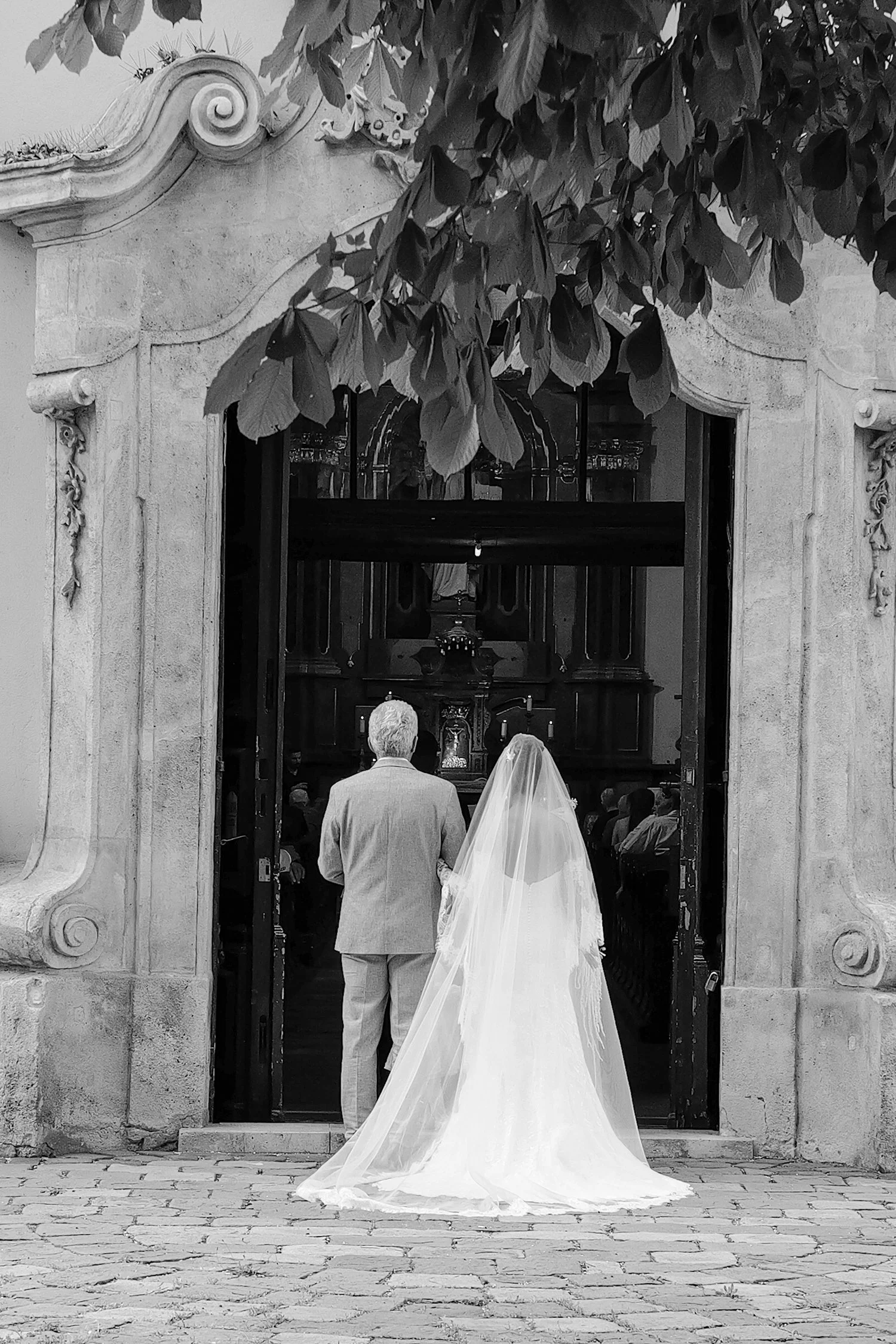 Une cérémonie de mariage à l'extérieur d'une ancienne entrée ou portail en pierre, avec un couple marié debout face à l'entrée, la mariée en robe blanche avec voile, le marié en costume clair, sous un arbre avec des feuilles abondantes.