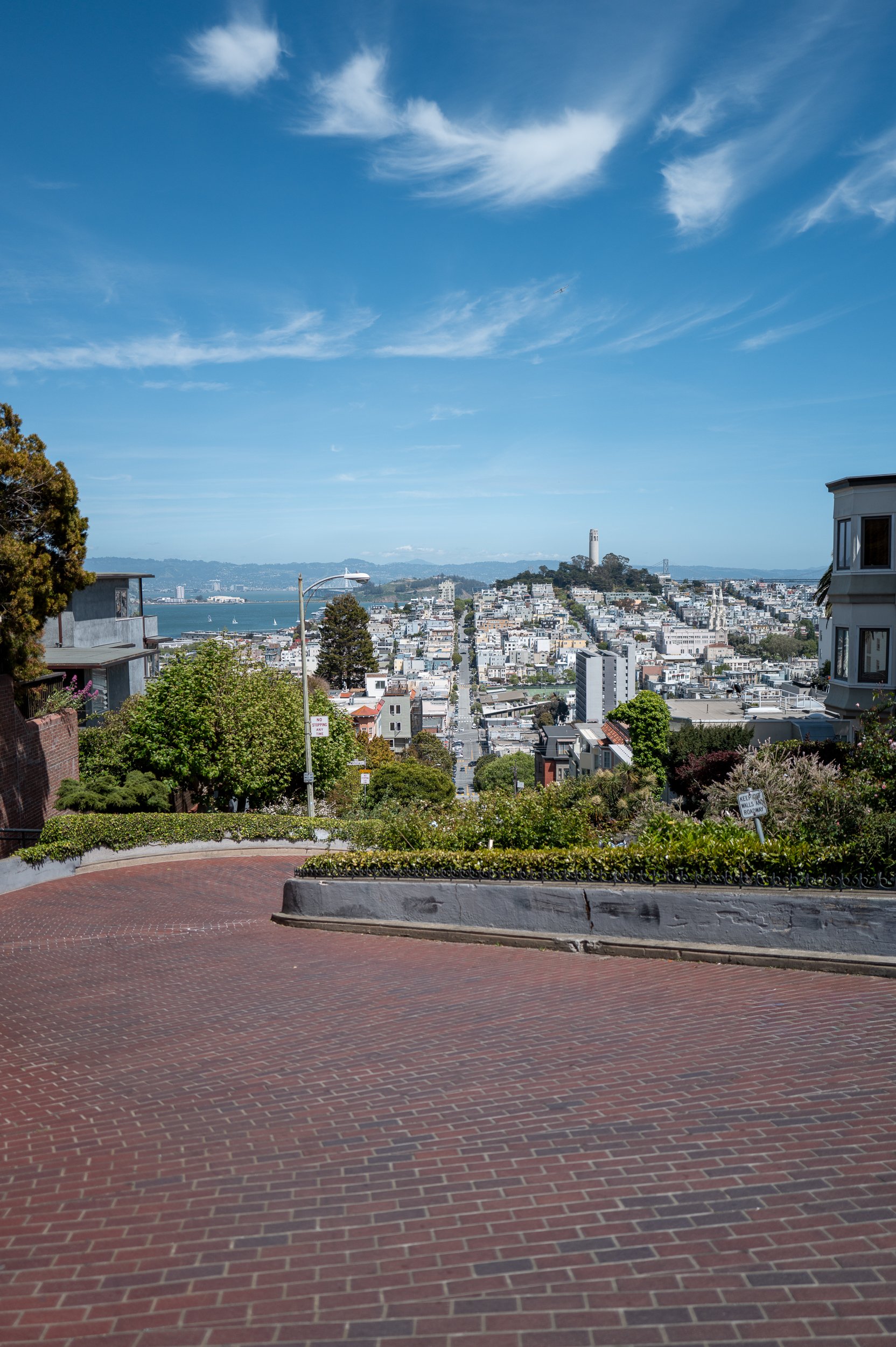 Vue d'une ville avec des bâtiments, une colline avec une tour, un ciel bleu avec des nuages, un peu de verdure et une surface pavée au premier plan.