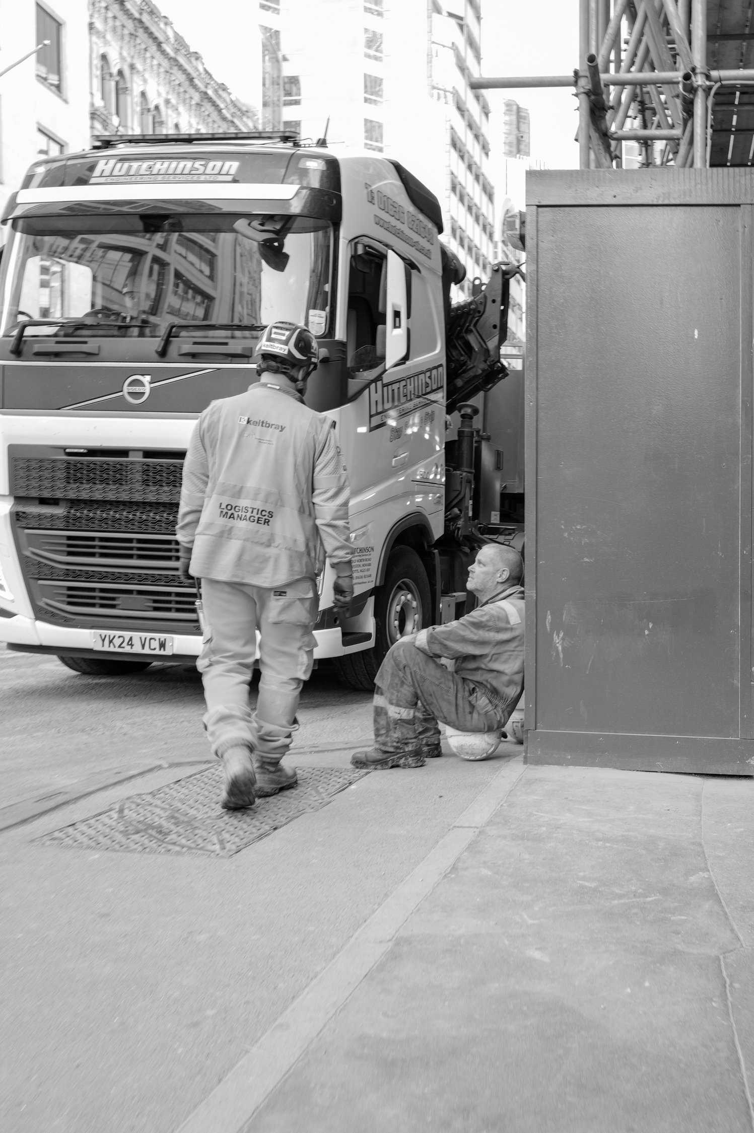 Deux hommes travaillant à côté d'un camion de logistique sur une rue urbaine avec des bâtiments en arrière-plan. L'un est debout, portant un gilet indiquant 'Logistics Manager', l'autre est assis, tous deux portant un casque de sécurité.