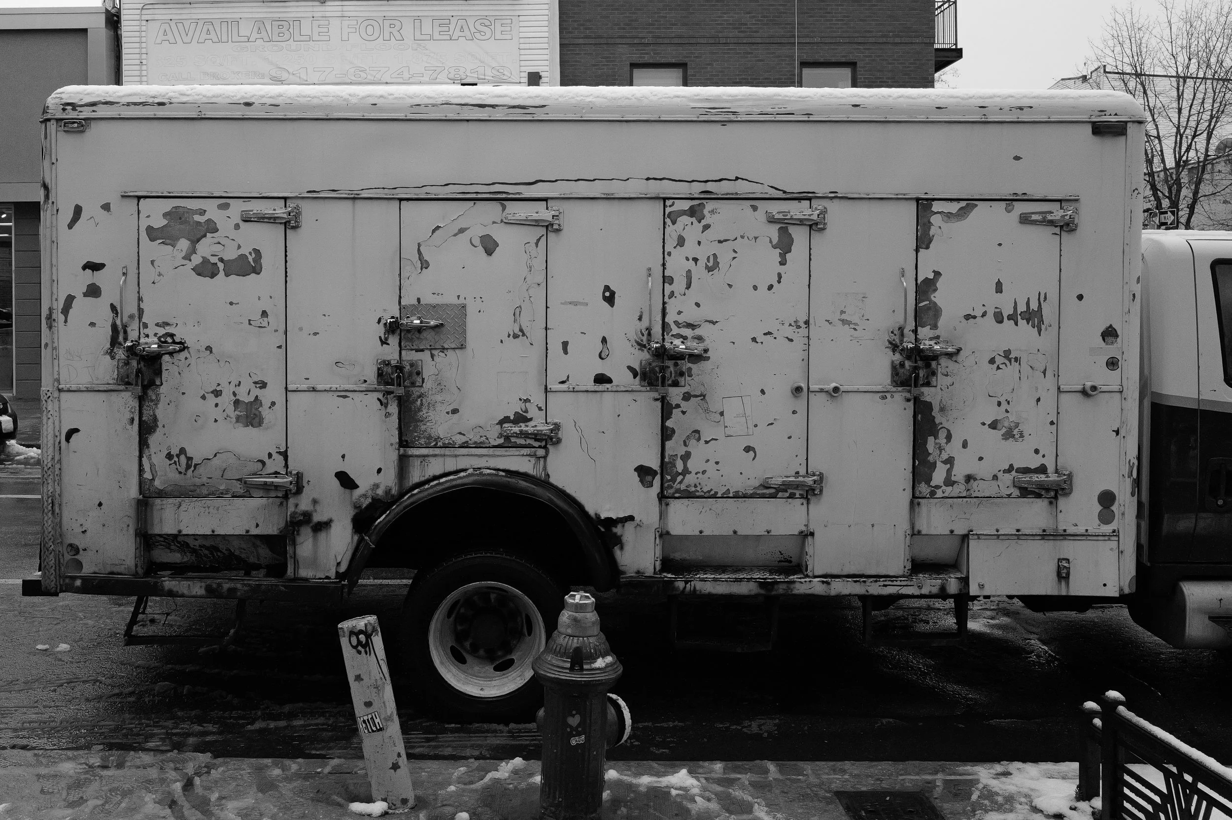 Camion de livraison abandonné avec une peinture écaillée et sale, stationné sur le trottoir avec un poteau d'interdiction, un feu de circulation et un sèche-mains, dans une zone urbaine. Bâtiments en arrière-plan avec affiches, y compris une panneau 