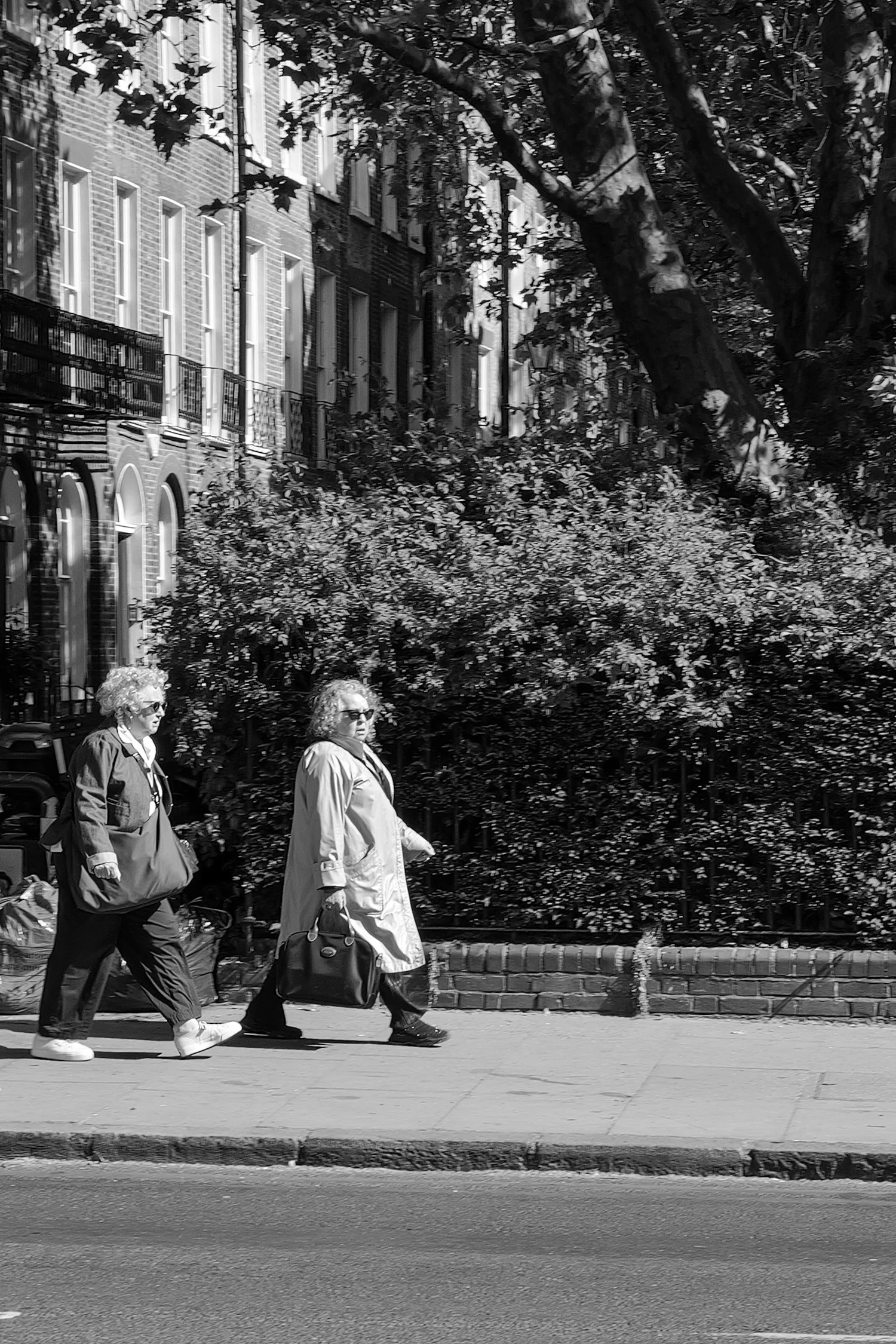 Trois femmes marchent le long d'un trottoir bordé d'arbres et de bâtiments résidentiels en briques, en noir et blanc.
