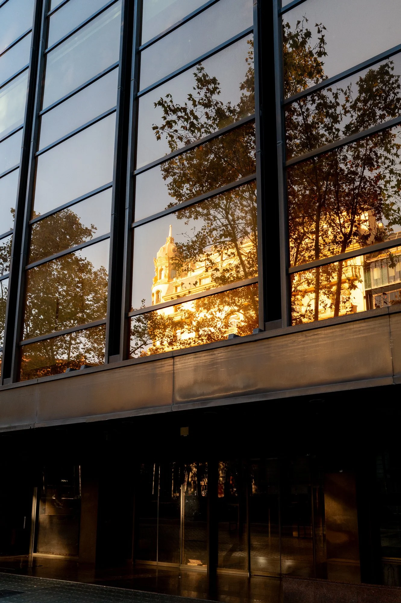 Reflet d'un bâtiment ancien dans les fenêtres d'un bâtiment moderne en verre, avec des arbres et une lumière dorée du coucher du soleil