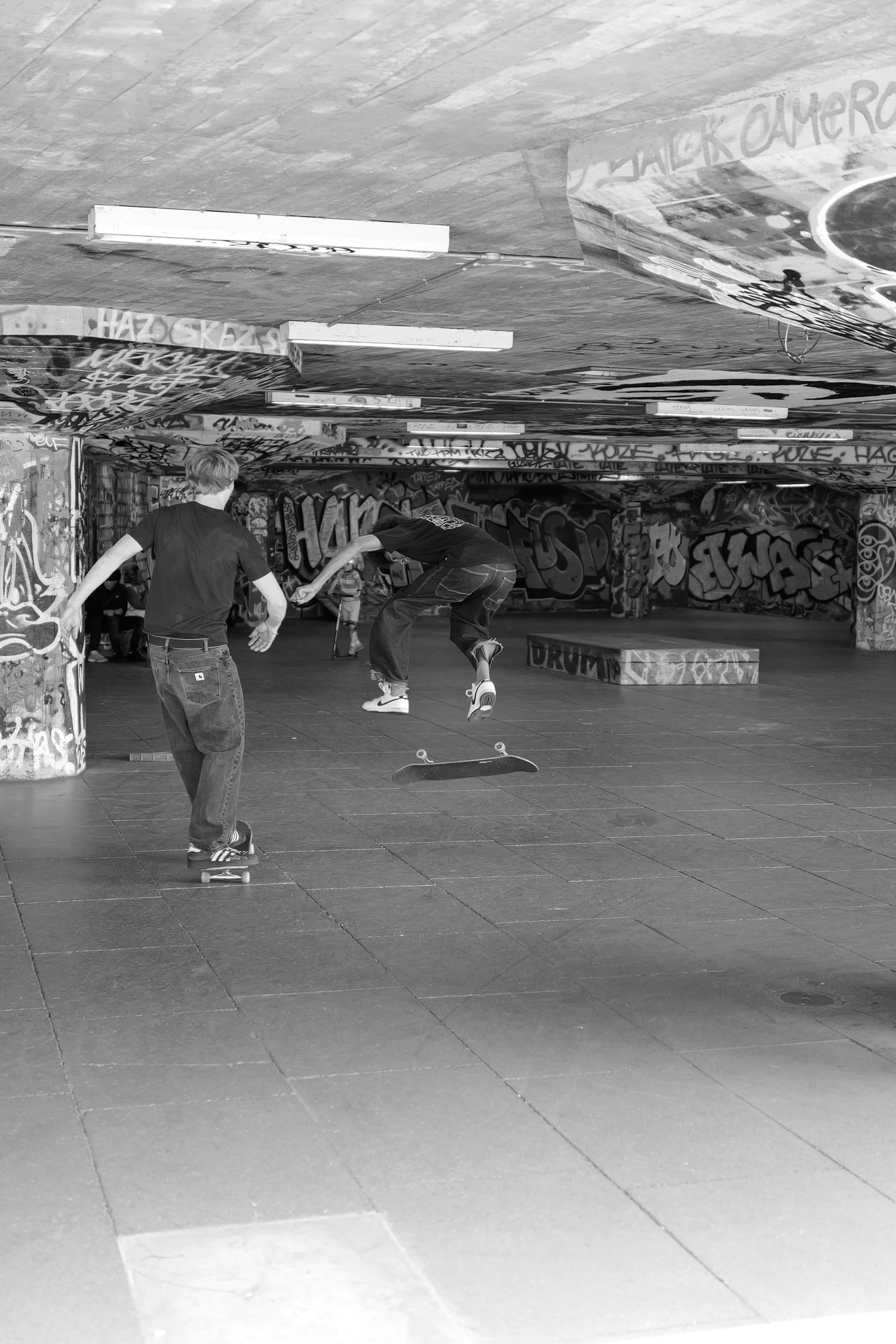 Deux jeunes faisant du skateboard dans un espace urbain sous un pont graffité.