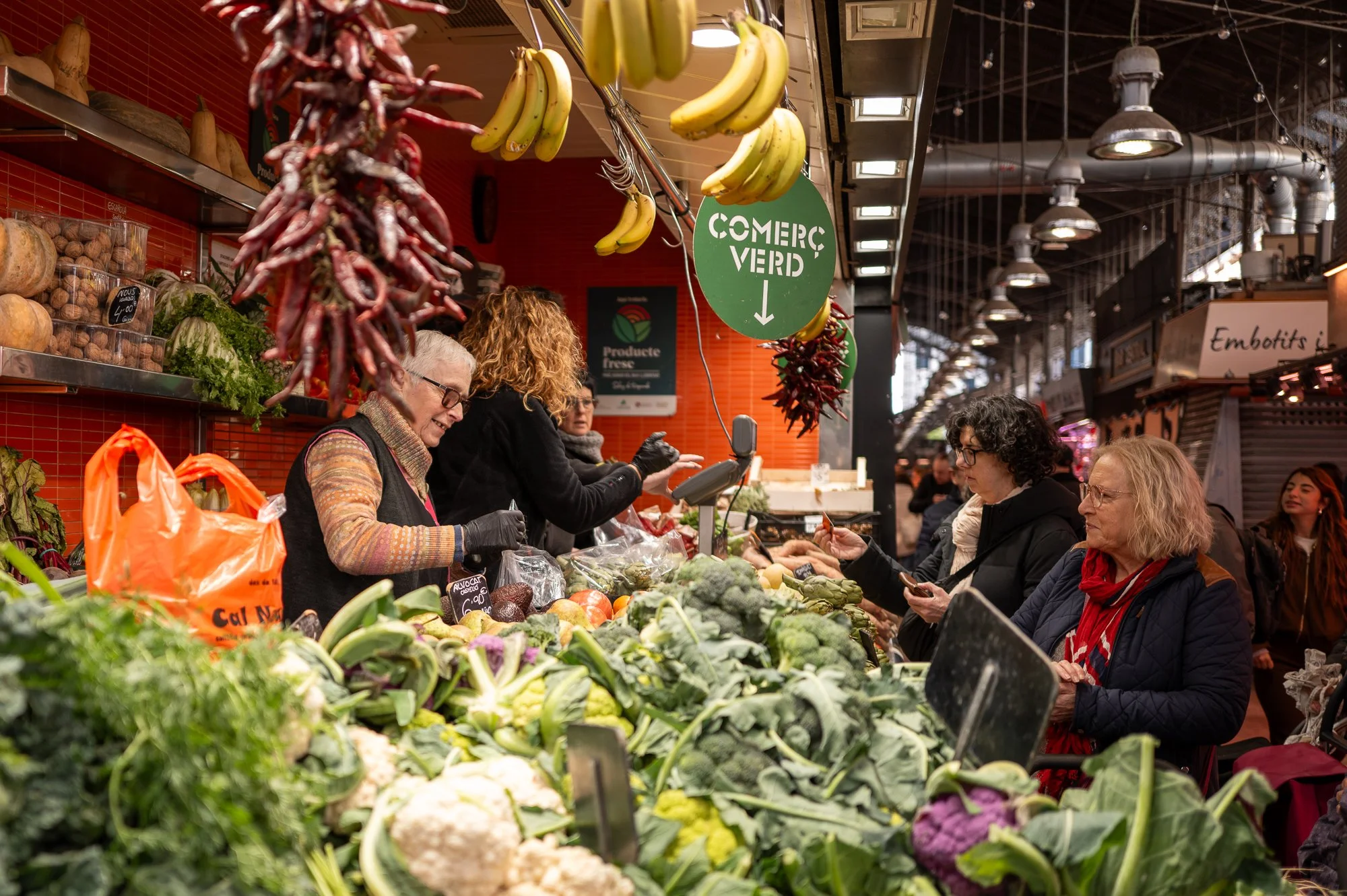 Marché couvert avec vendeurs vendant des légumes et des fruits biologiques, clients faisant leurs achats, panneaux indiquant 'Comerc Verd'.