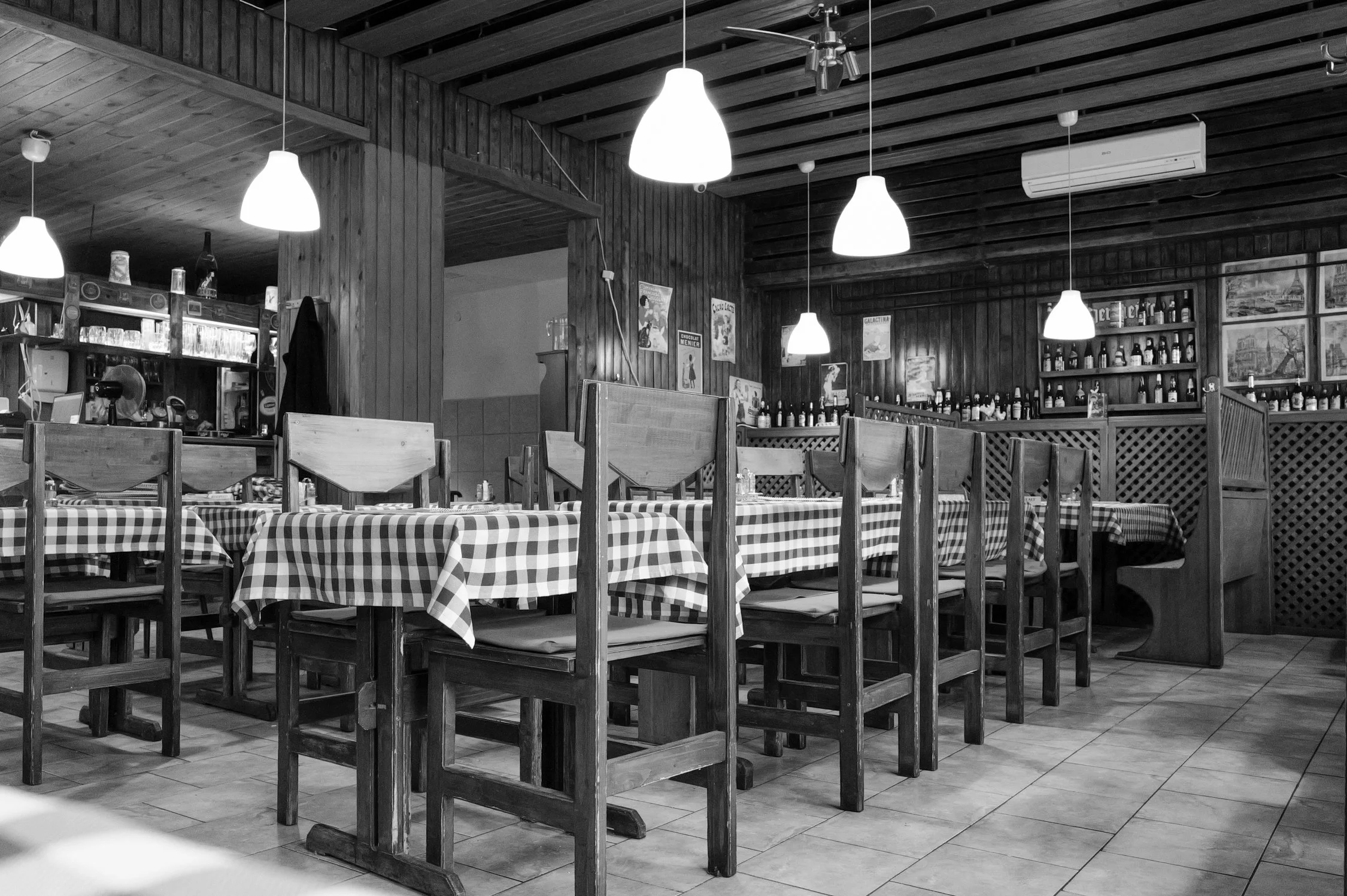 Une salle de restaurant en bois avec des tables à nappes à carreaux et des chaises en bois. Il y a des lampes suspendues au plafond et des posters sur les murs, avec un mur en arrière-plan où sont exposées des bouteilles et des images.