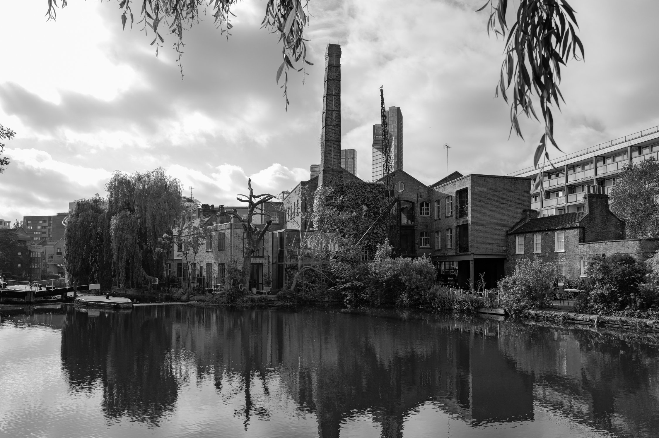 Une scène urbaine en noir et blanc avec des bâtiments résidentiels et industriels, un canal ou étang au premier plan, des arbres et un ciel nuageux.