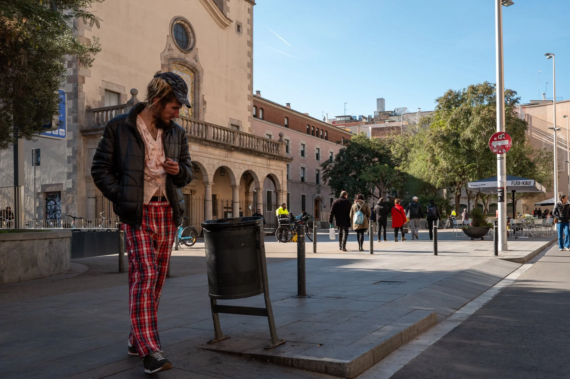 Un homme en pyjama à carreaux rouges et blancs, portant une veste en cuir noire et un chapeau, regarde son téléphone en dehors d'un bâtiment historique, avec plusieurs personnes marchant dans la rue.